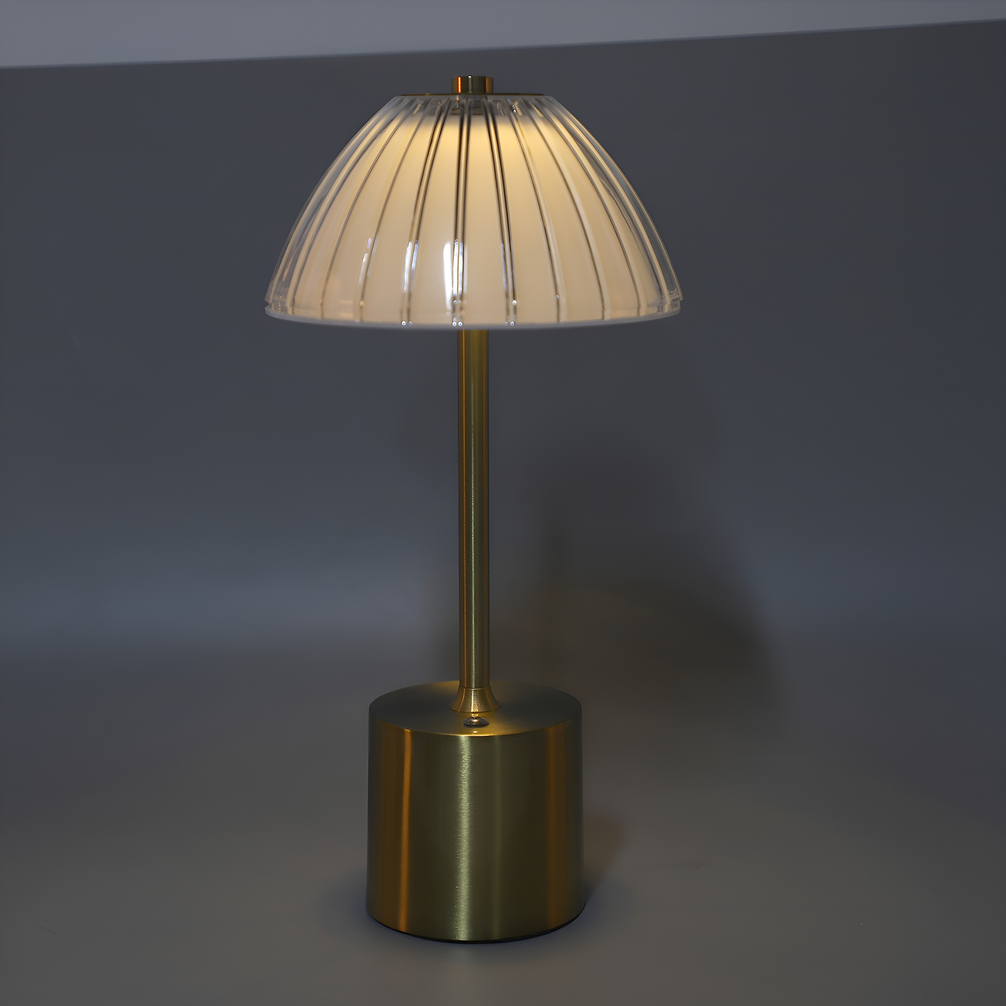 Eine goldfarbene Tischlampe mit einem klaren, gerippten Lampenschirm. Die Lampe steht auf grauem Grund.