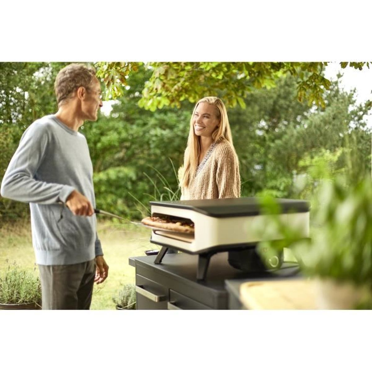 Een stel bereidt pizza op een pizza-oven in de buitenlucht, met groen op de achtergrond.