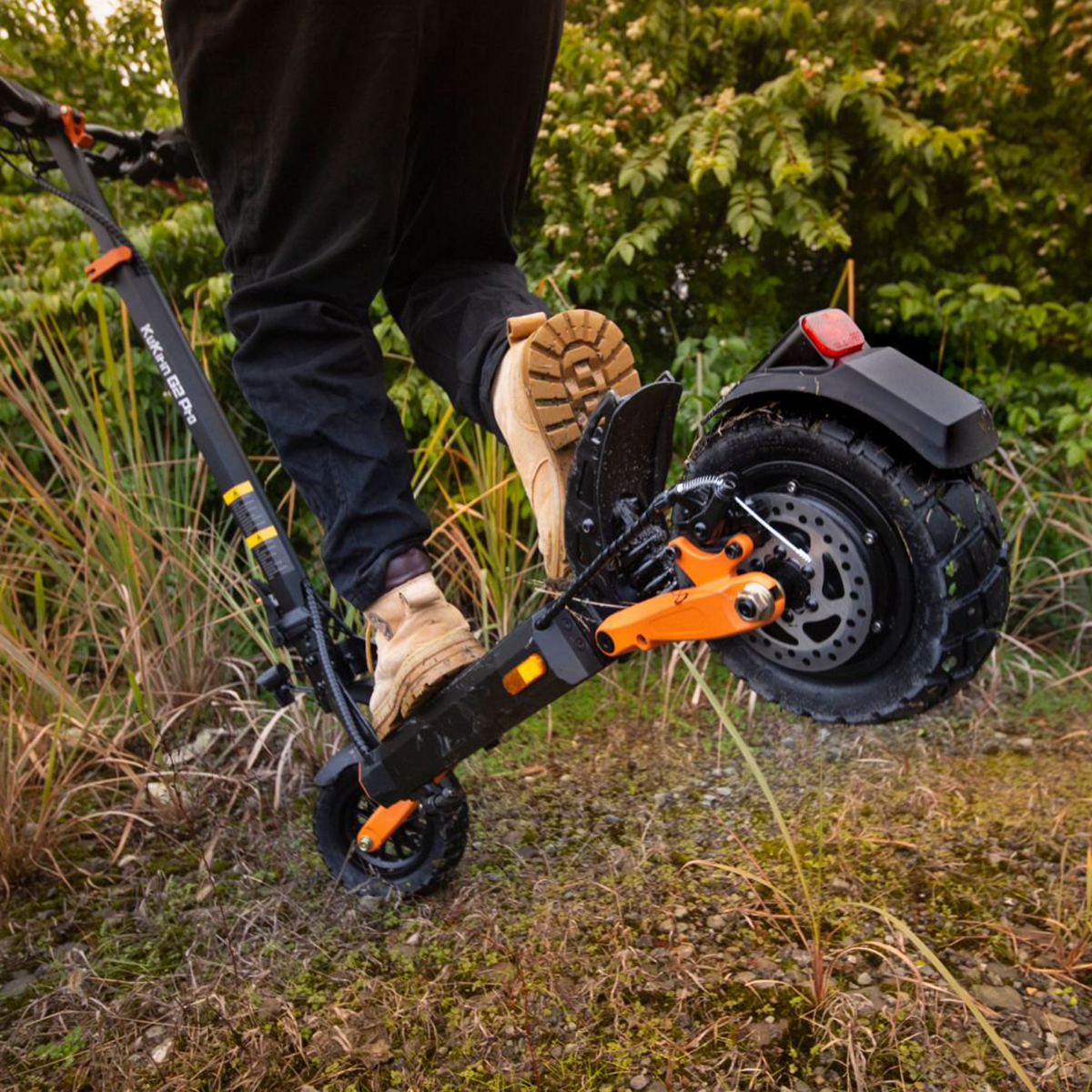 Persona montando un patinete eléctrico negro y naranja en un camino cubierto de hierba con plantas alrededor.