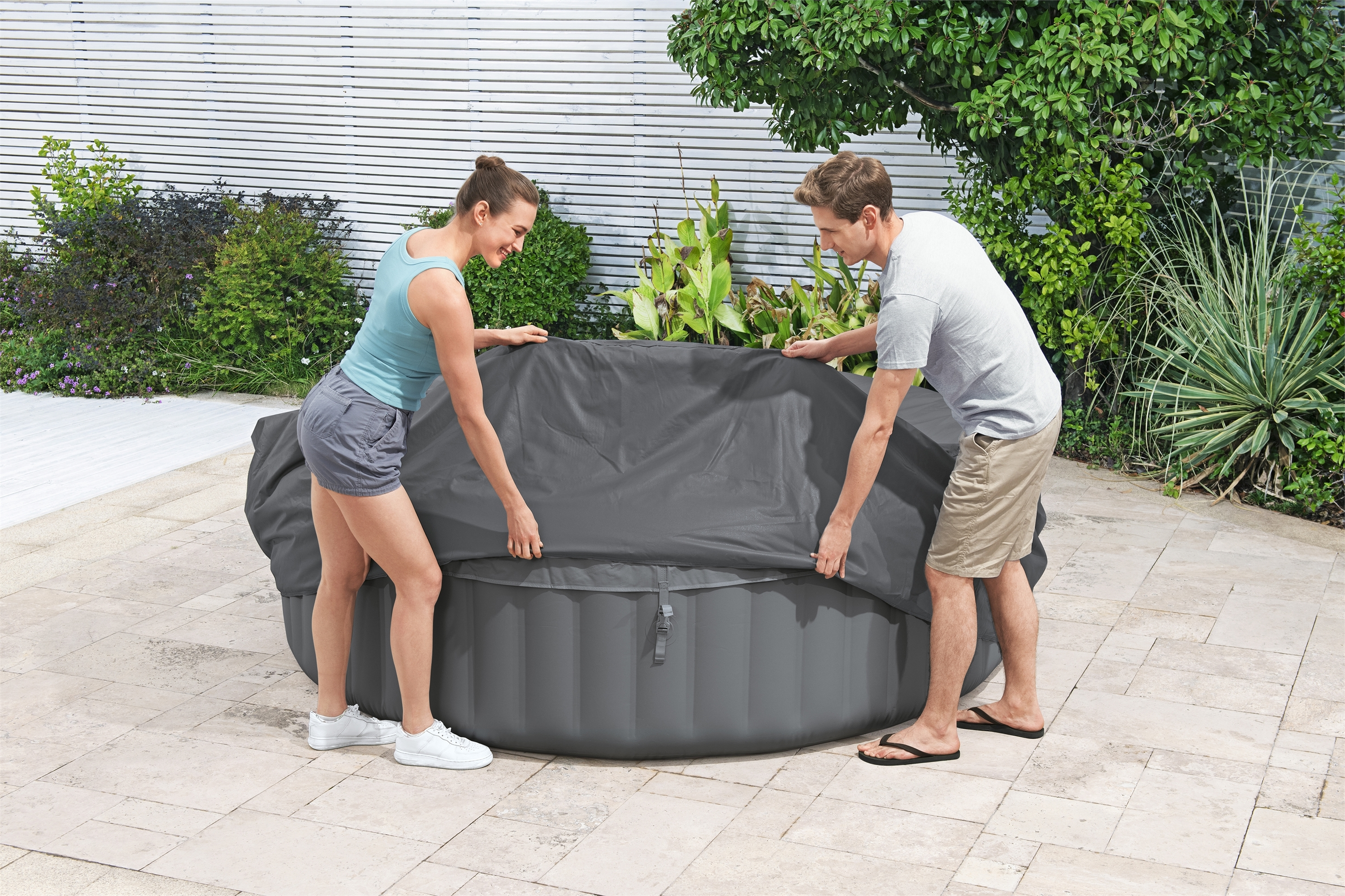 Ein Mann und eine Frau bedecken einen runden, grauen Whirlpool mit einer dunkelgrauen Abdeckung.