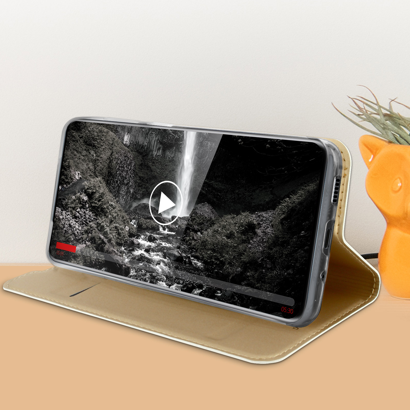 Custodia per telefono con video in riproduzione, una scena di cascata. Un piccolo vaso a forma di gatto arancione si trova vicino.