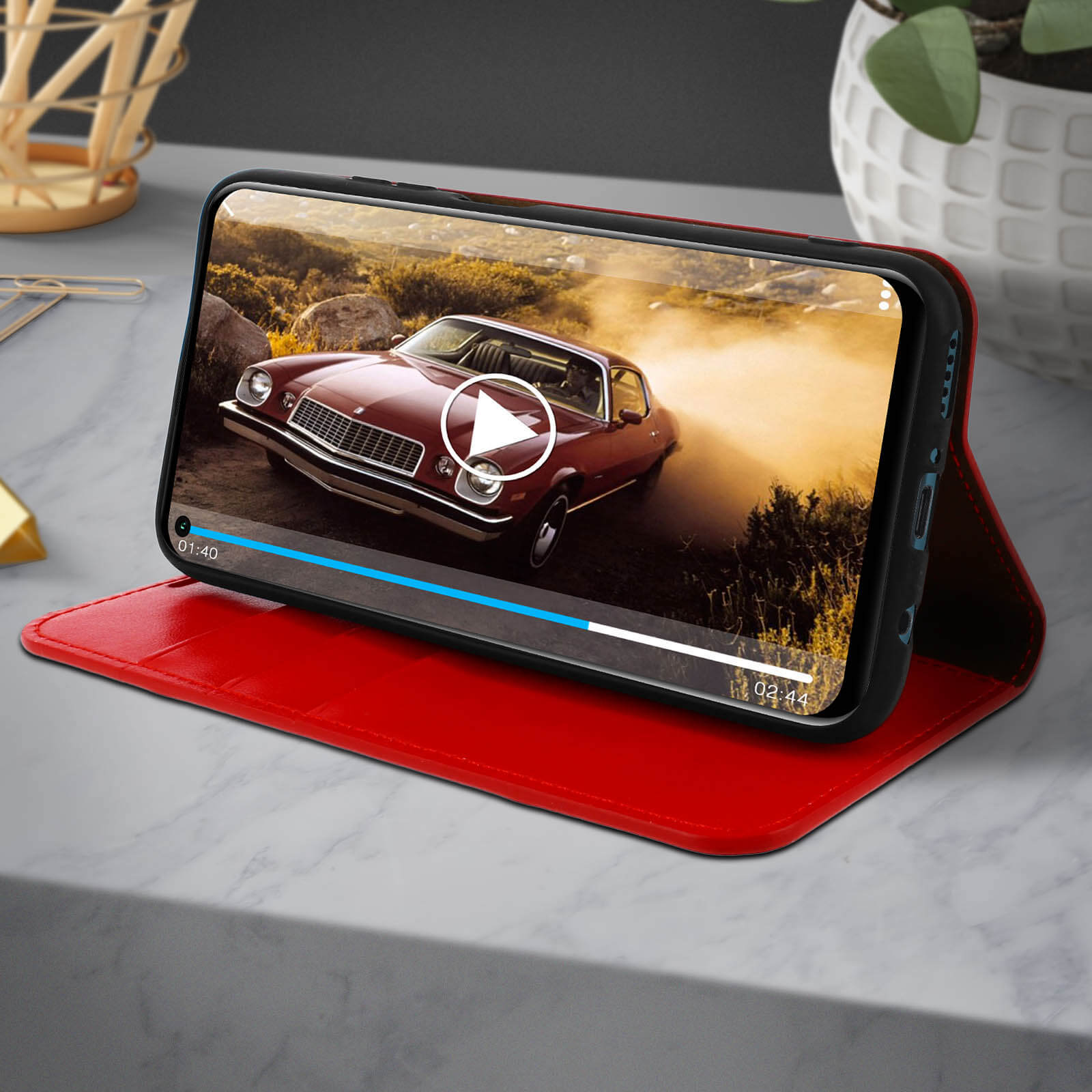 Telefono in custodia rossa che mostra video di un'auto. Superficie in marmo. Sfondo: auto sfocata.