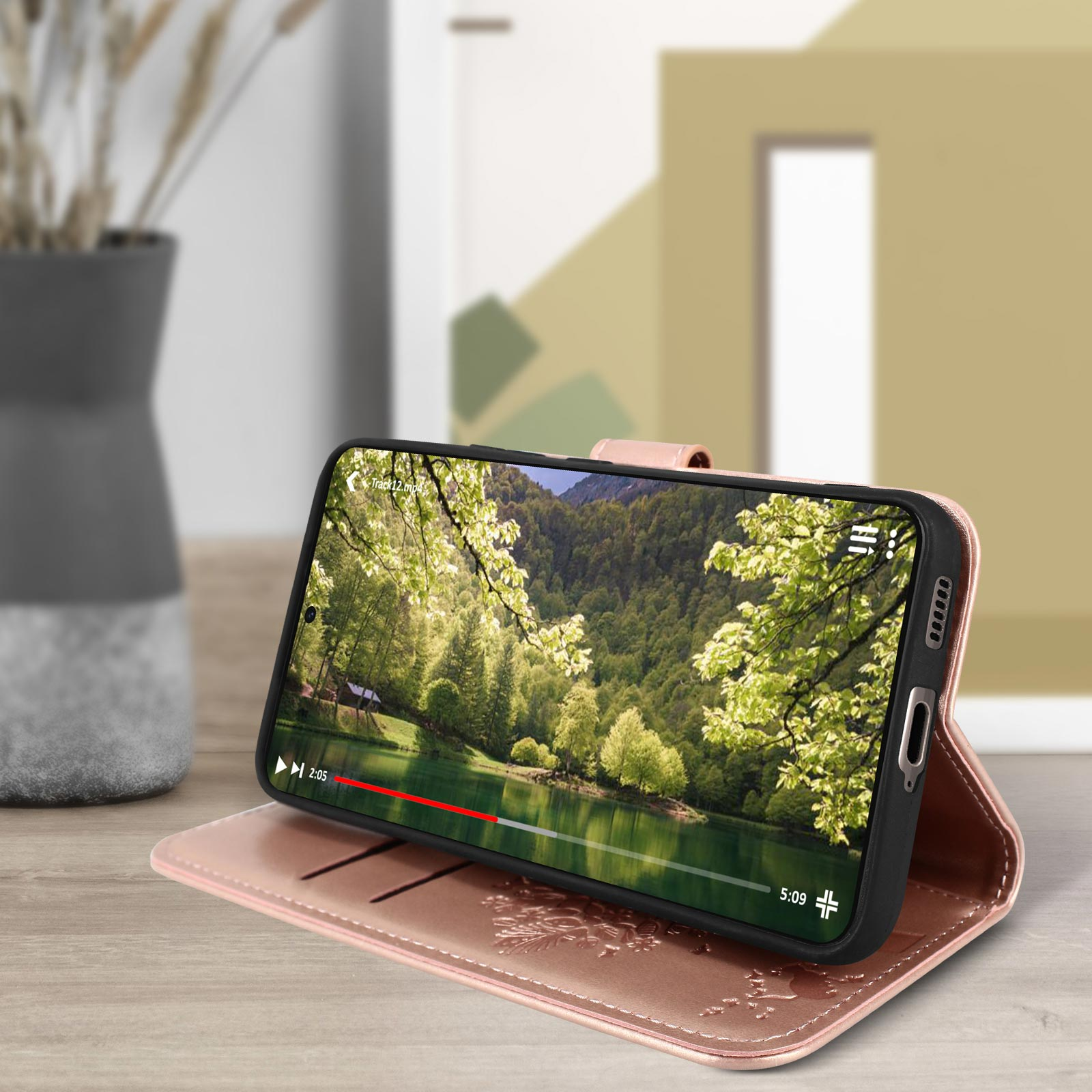 Telefono in custodia rosa, riproduce video, su un tavolo. Sfondo: alberi verdi e scena del lago.