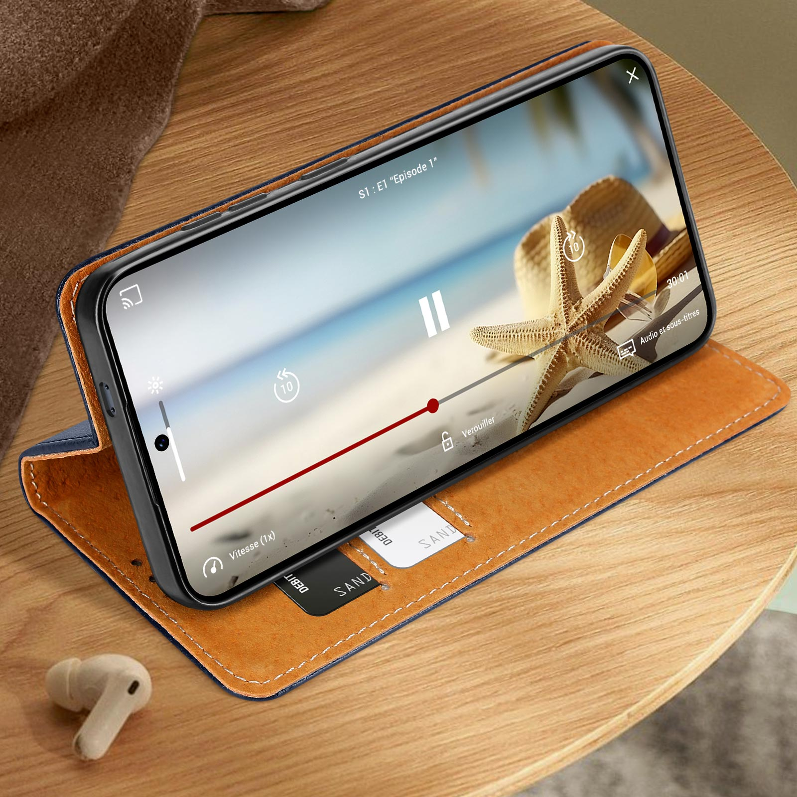 Geöffnete Handyhülle auf einem Holztisch. Telefon spielt Video ab. Kopfhörer auf dem Tisch. Karten sichtbar.