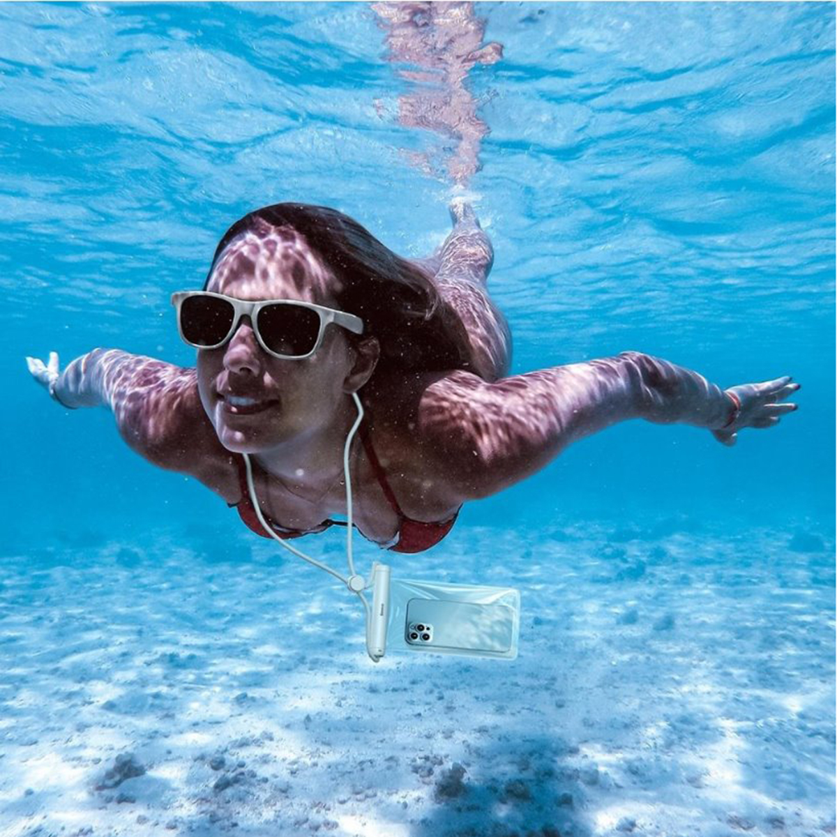 Donna che nuota sott'acqua con occhiali da sole, bikini rosso e telefono in custodia impermeabile.
