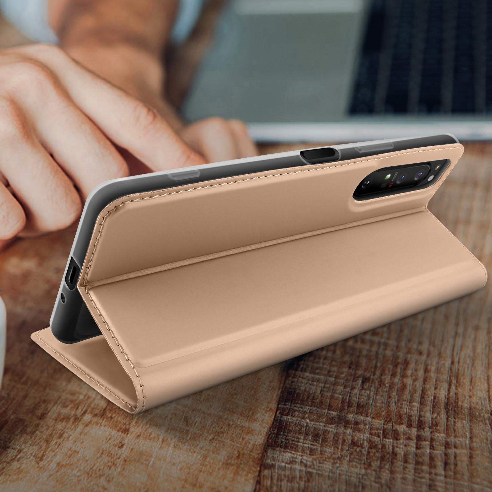 Un telefono in una custodia in pelle beige è su una superficie di legno, parzialmente aperto. C'è una mano vicina.