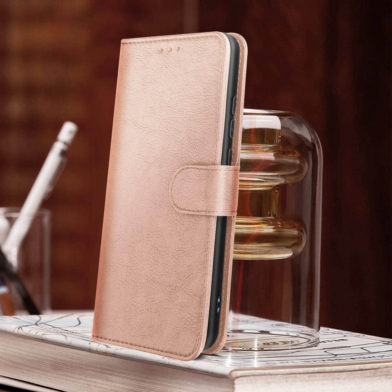 Custodia rosa per telefono, aperta, su un libro. Un vaso di vetro sullo sfondo, con una penna a sinistra.