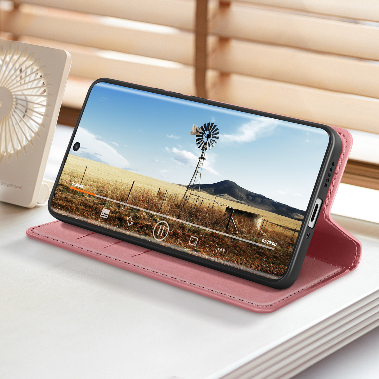 Telefono in custodia rosa, aperto su superficie bianca, mostra video con mulino a vento sullo sfondo.