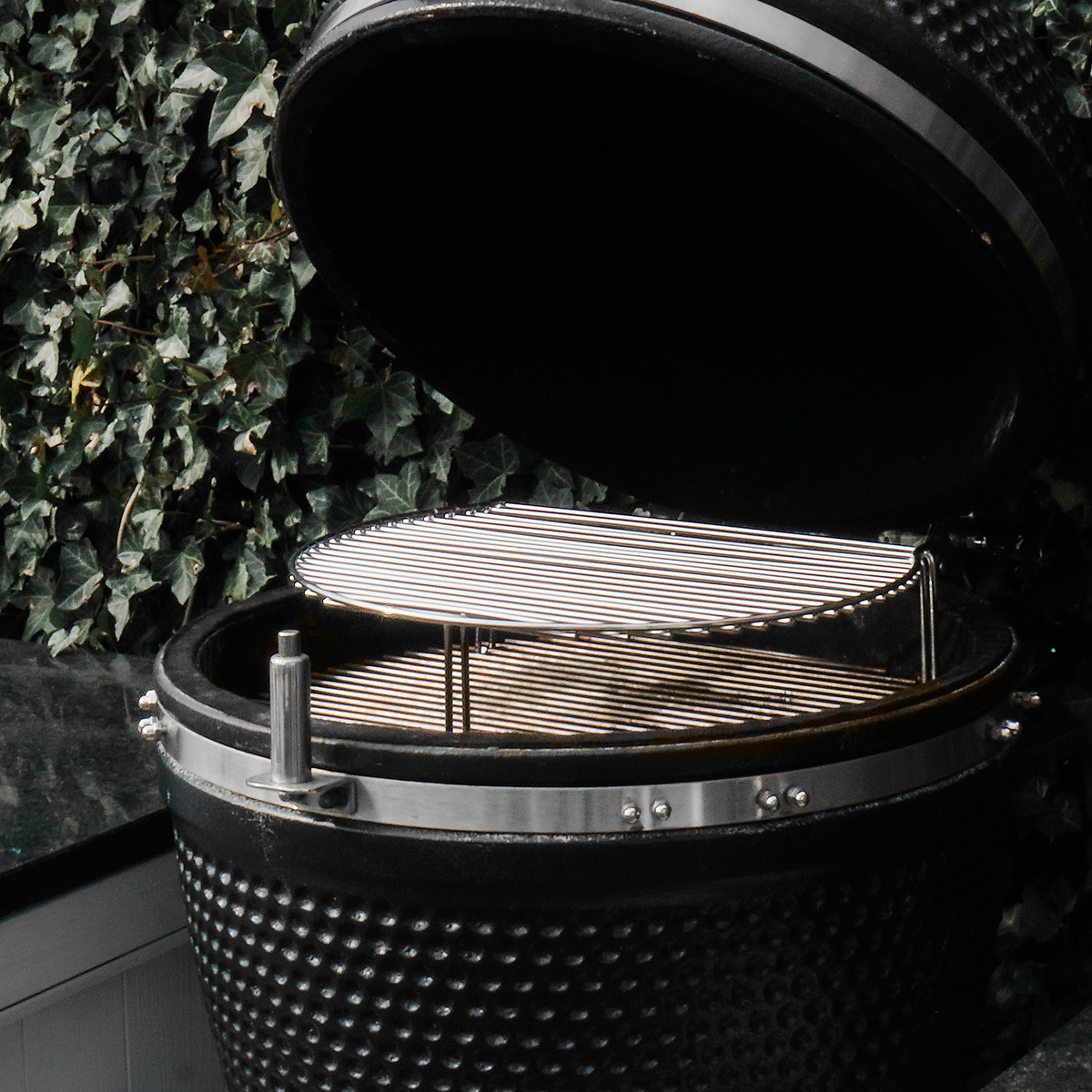 Black grill with the lid open and a metal grill inside. Green leaves are in the background.