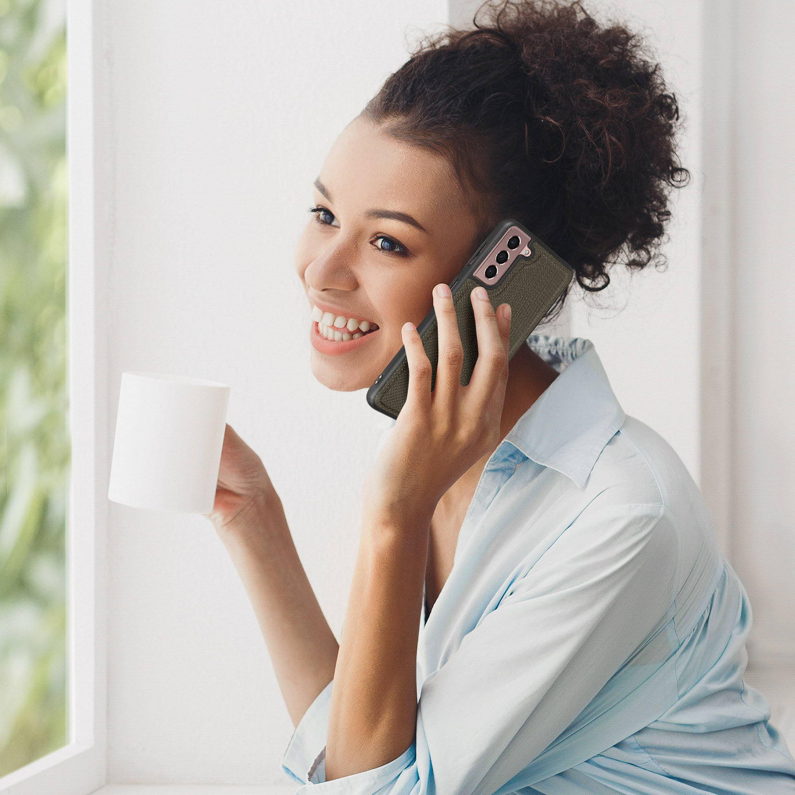 Donna al telefono con telefono con custodia verde, sorridente. Tazza bianca nella mano sinistra. Finestra.