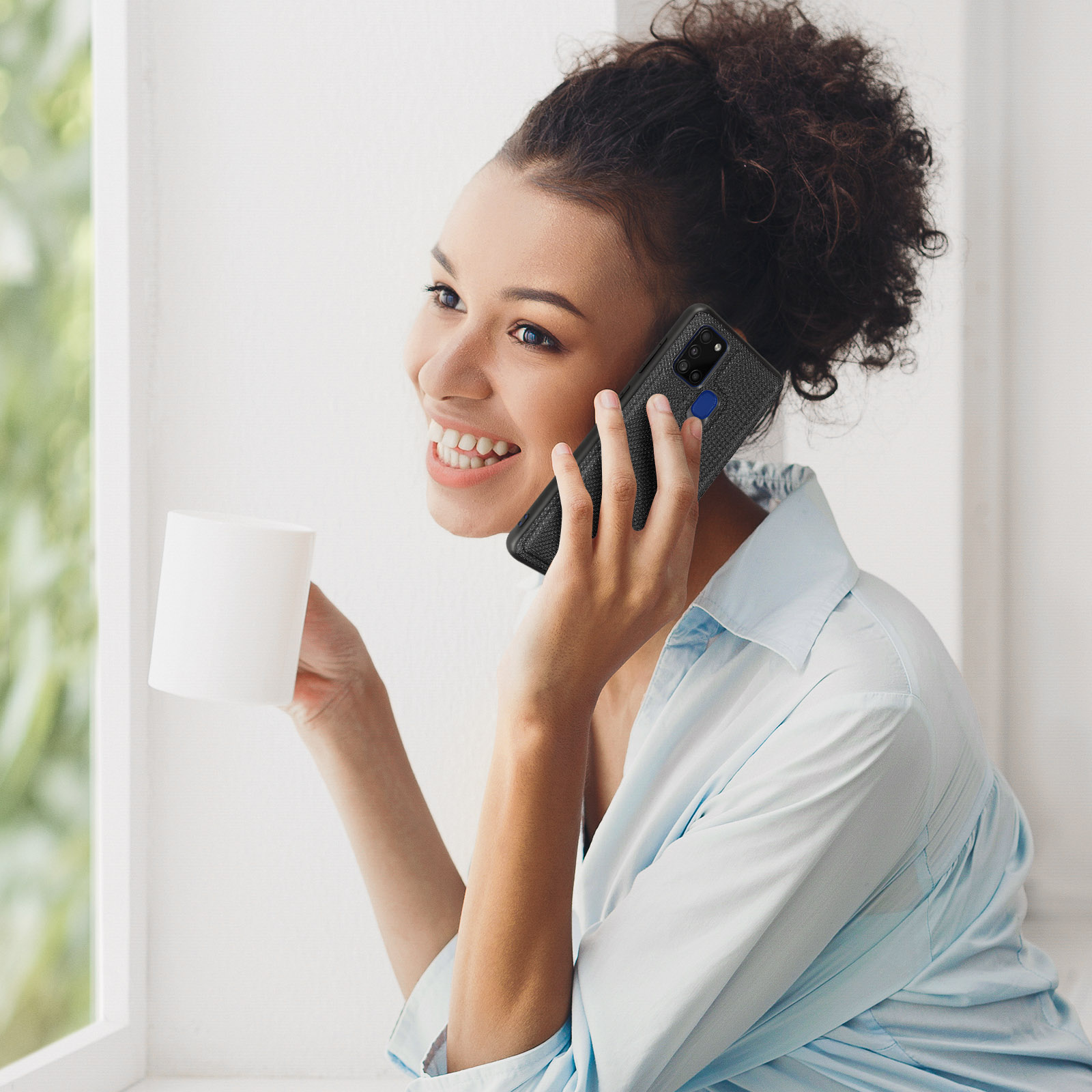 Donna sorridente al telefono, tazza bianca. Luce solare vicino alla finestra. Custodia nera visibile.