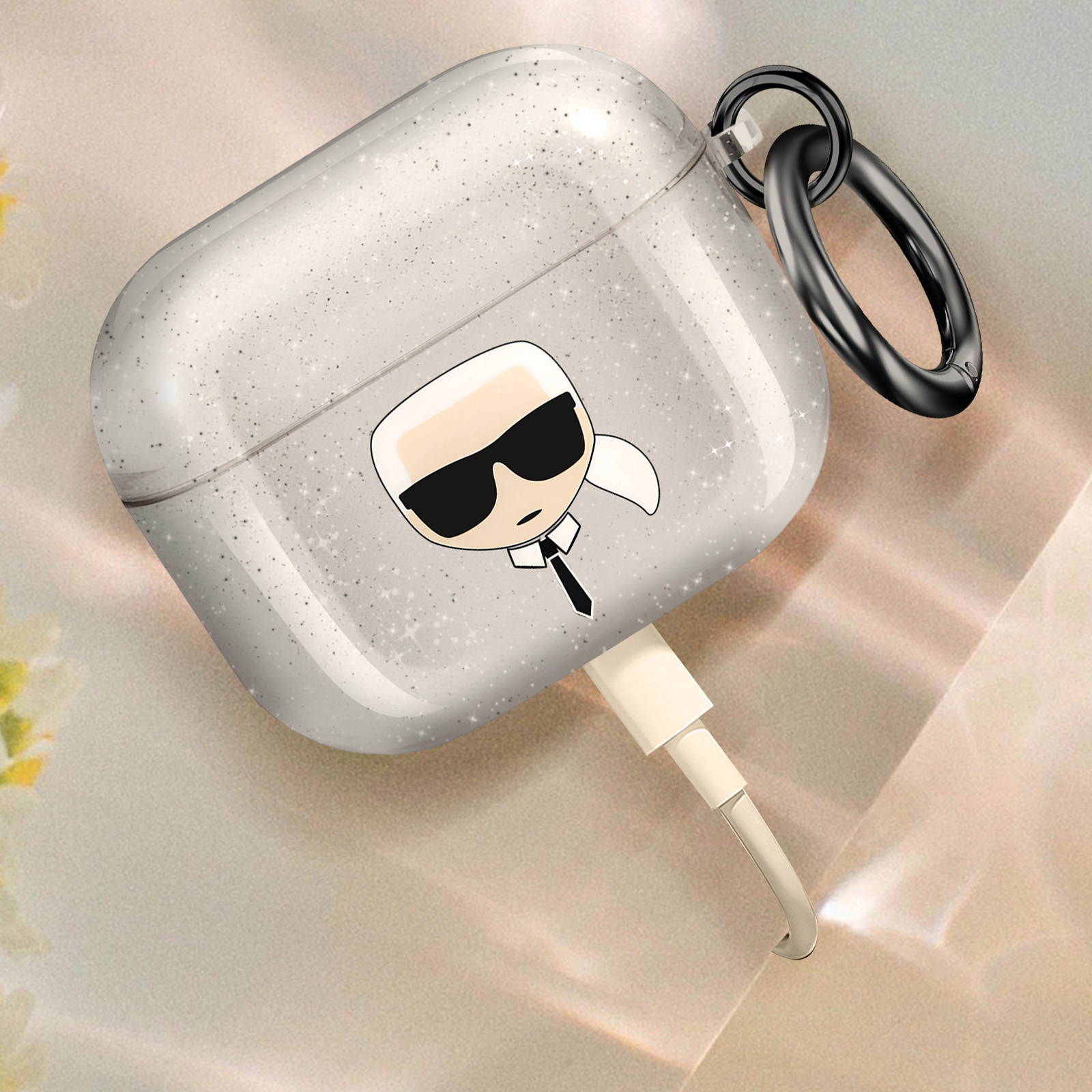 Custodia AirPods glitterata, design Karl Lagerfeld. Un cavo di ricarica è collegato. Sfondo bianco.