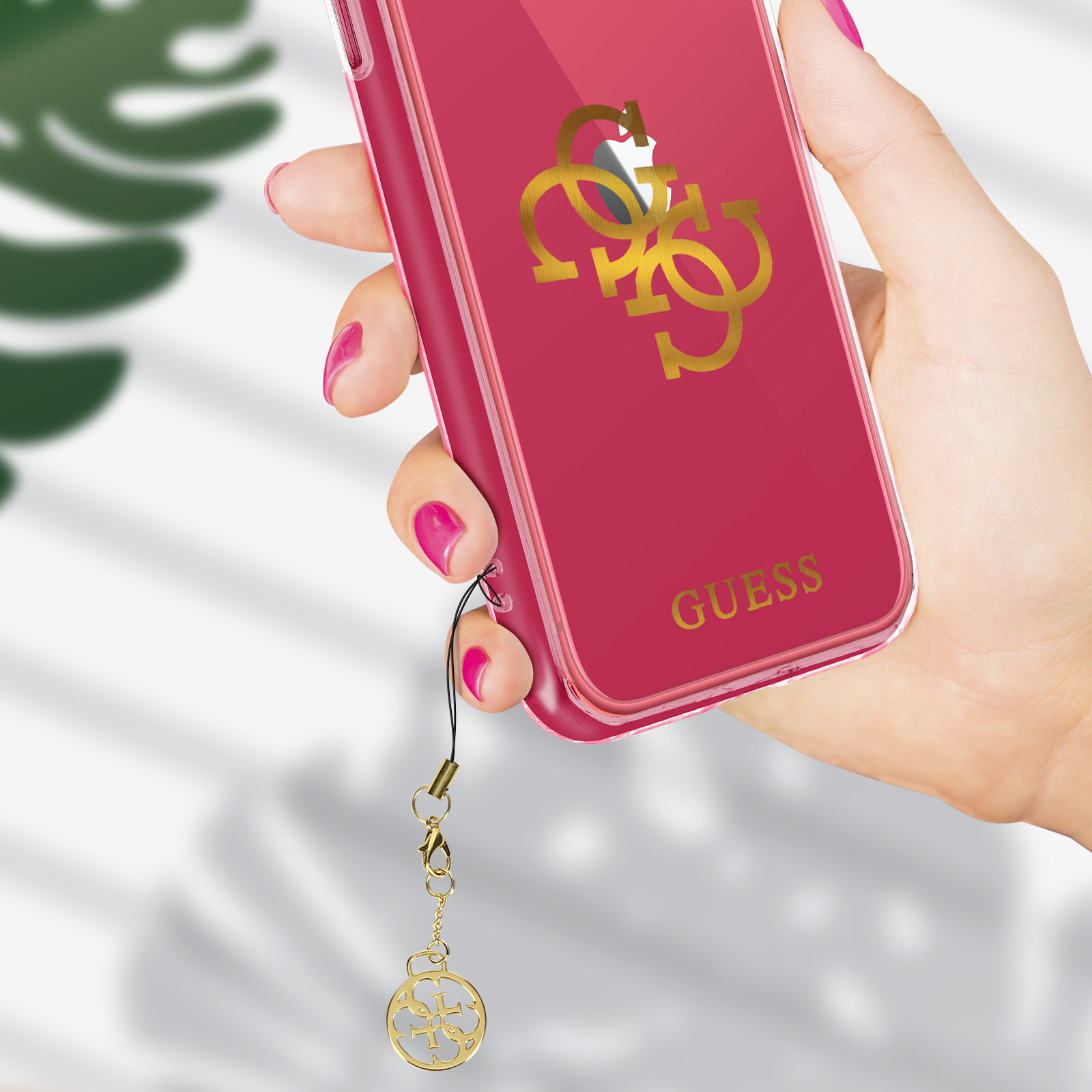 Hand hält Telefon mit roter GUESS-Hülle. Die Hülle hat ein goldenes Logo und einen Anhänger.