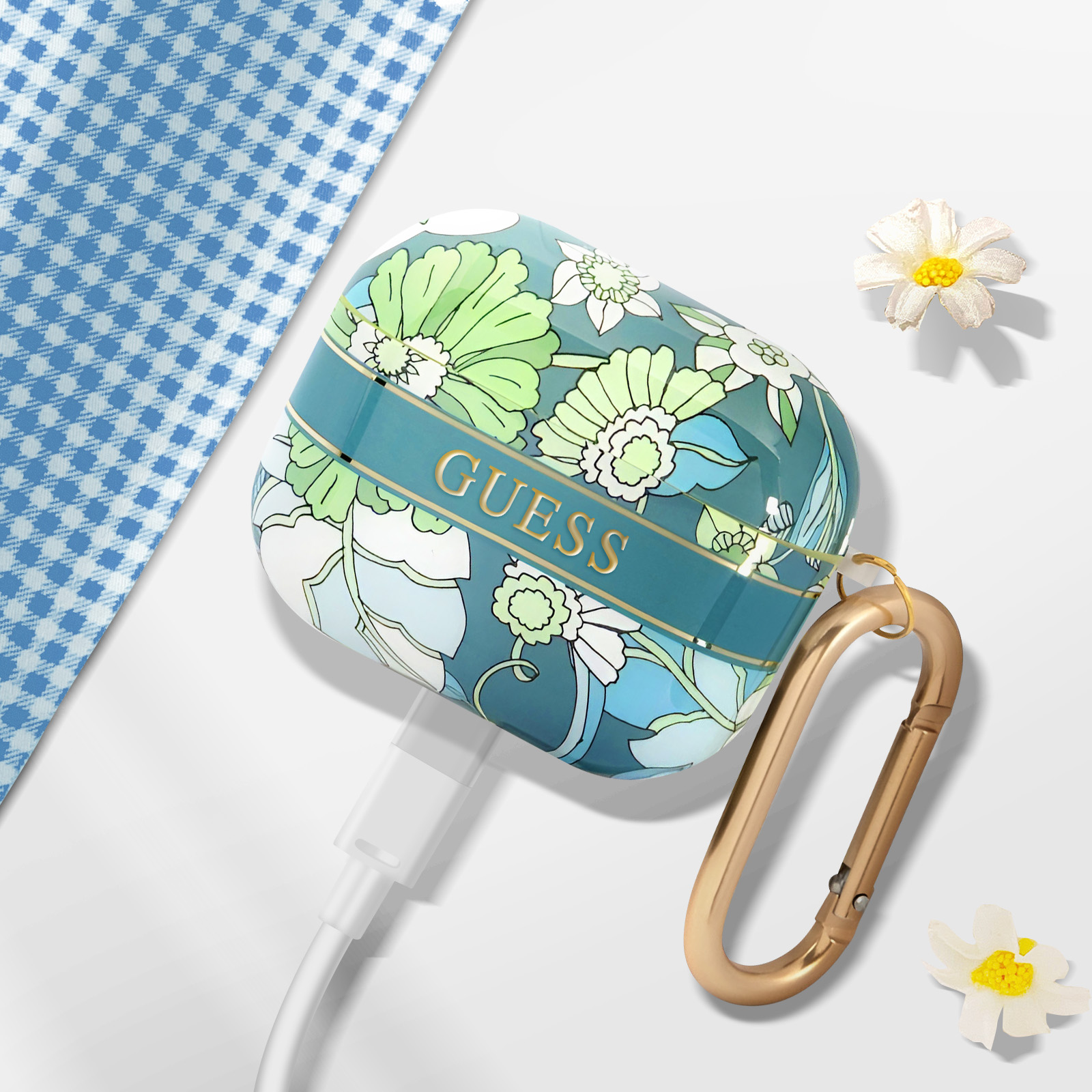 Una funda Guess para AirPods con un estampado floral, un cierre dorado y un cable de carga. Un paño a cuadros azul es visible.