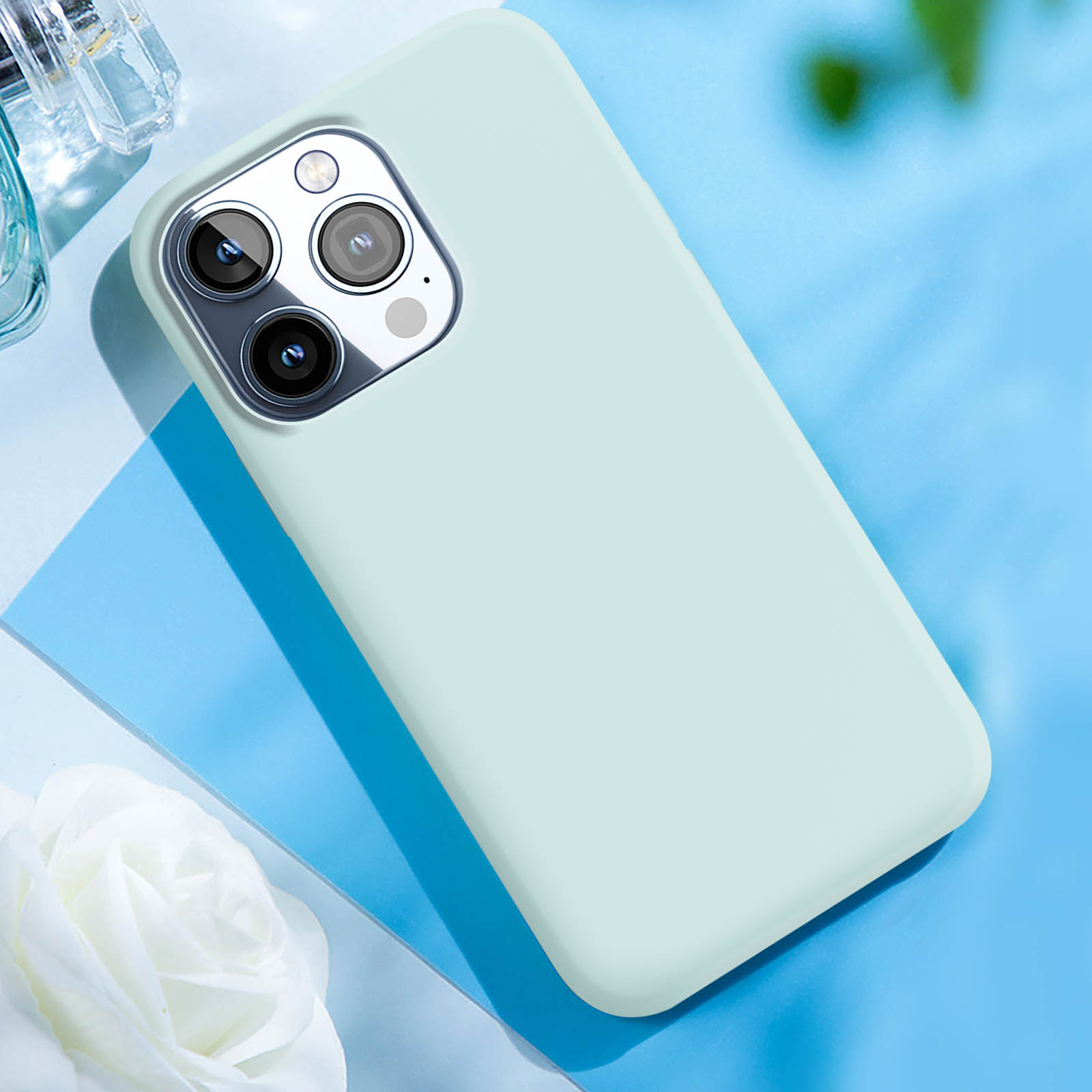 Custodia per telefono verde chiaro su una superficie blu. Sono visibili un fiore bianco e un flacone di profumo.