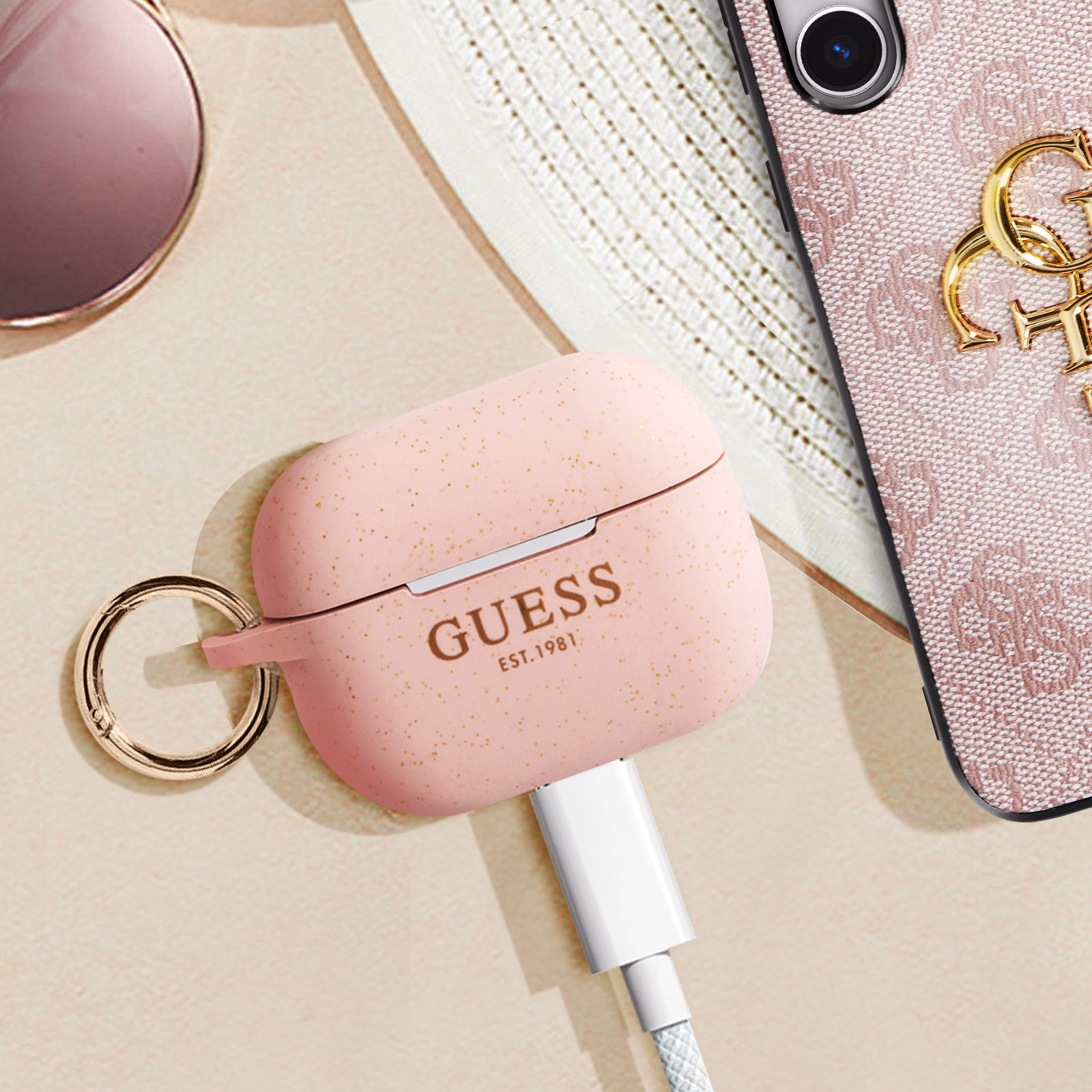 Funda rosa para AirPods con anillo dorado y marca Guess. Cable de carga blanco.