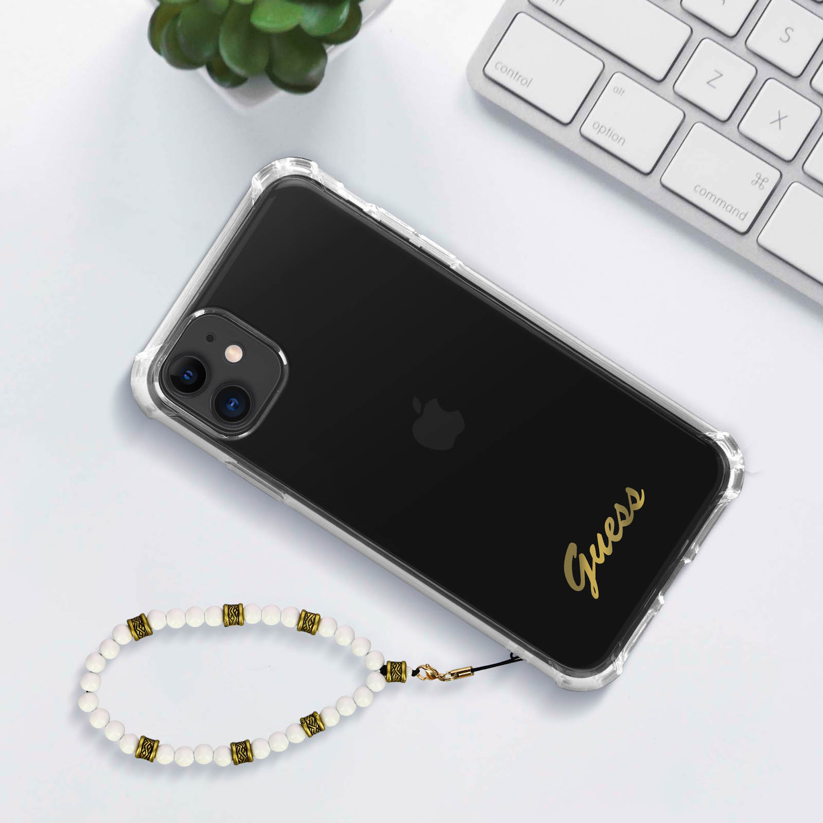 Custodia trasparente per telefono con logo Guess e cinturino da polso con perline bianche e dorate. Il telefono è su una superficie bianca.
