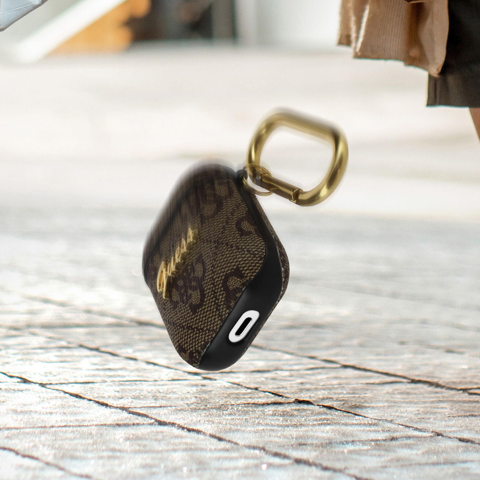 Una custodia marrone Guess per AirPods con un portachiavi dorato. Lo sfondo è sfocato.