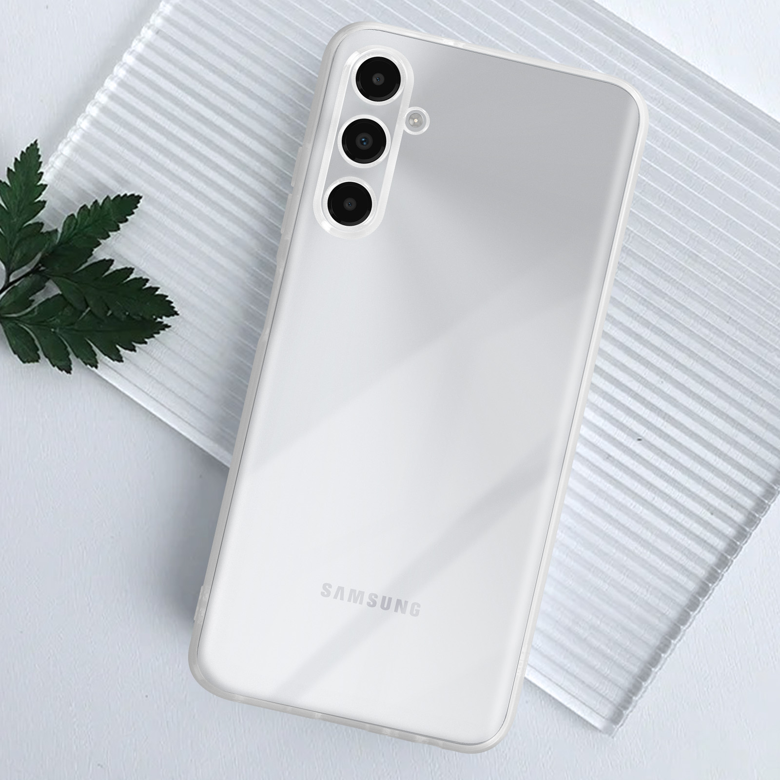 Telefono bianco con tre fotocamere su una superficie trasparente con una pianta verde. Il logo Samsung è visibile.