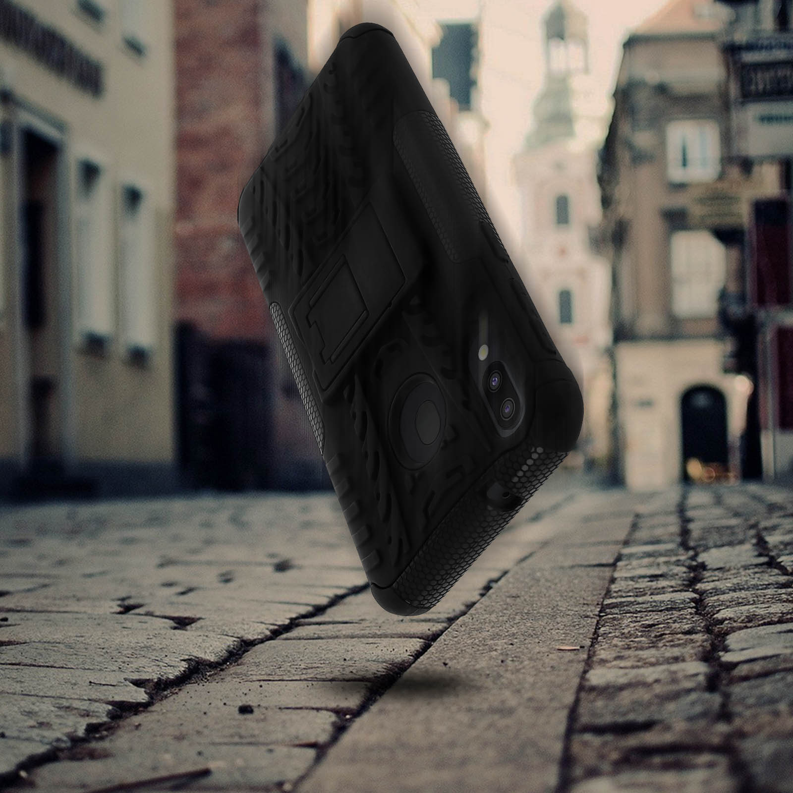 Custodia nera per telefono che levita sopra la strada acciottolata, con edifici sullo sfondo.