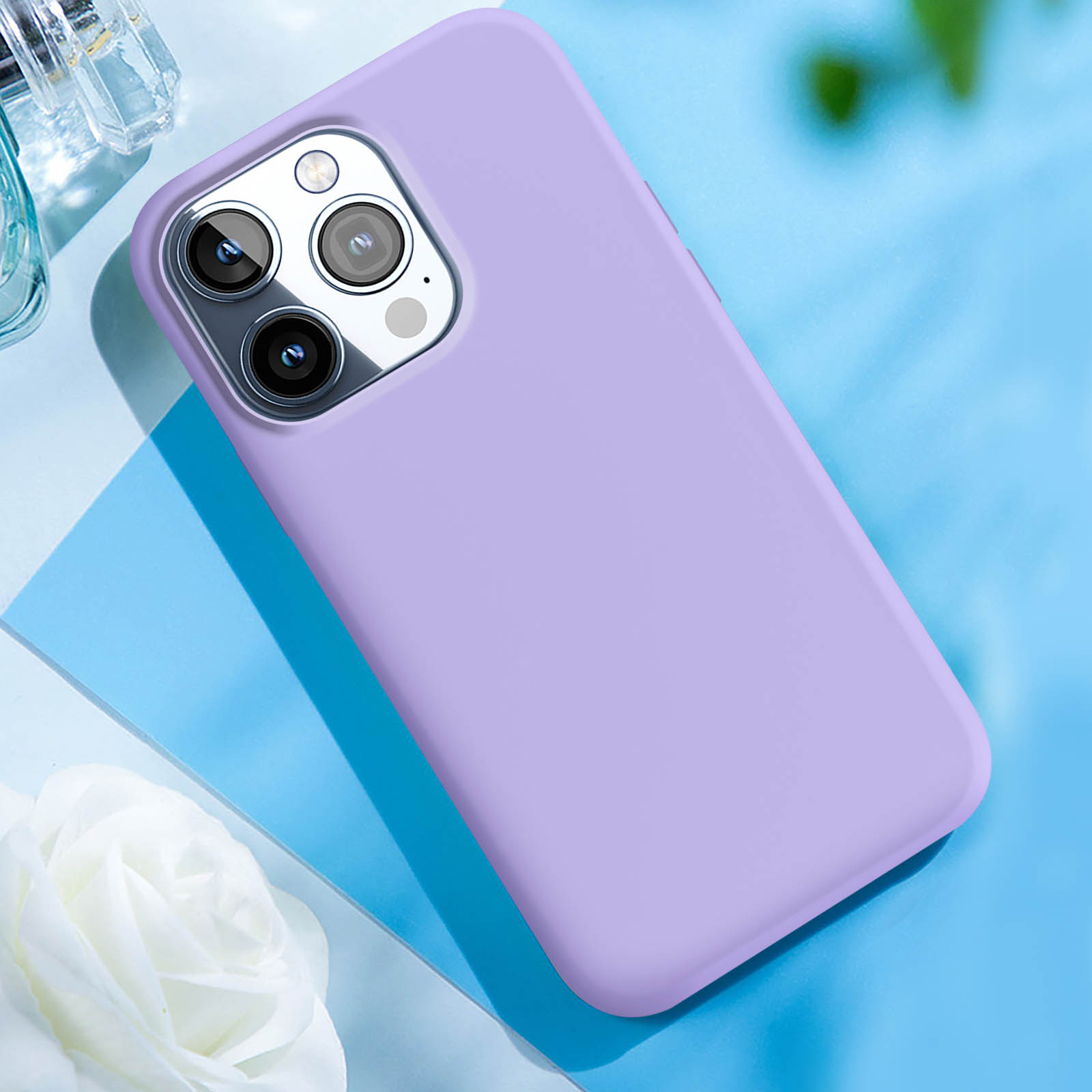 Una custodia per telefono viola su una superficie blu con un fiore bianco e una bottiglia di profumo. La custodia è a destra.