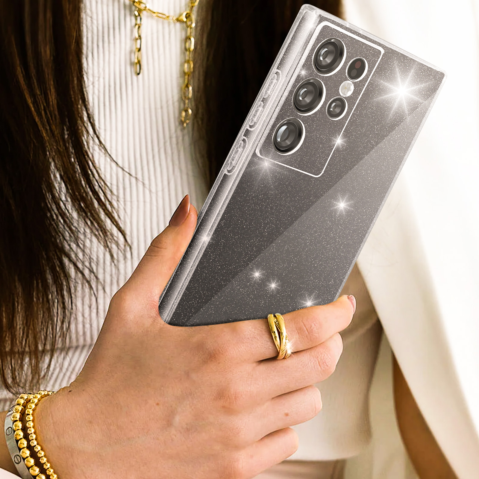 Una mano che tiene un telefono in una custodia glitterata. Gioielli d'oro sulla mano. Sfondo sfocato.