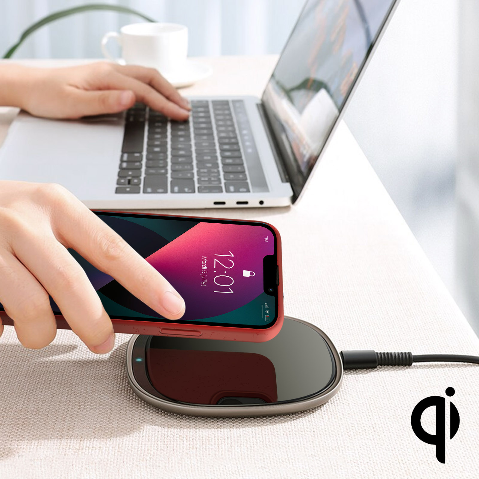 Una persona posiziona un telefono rosso su un caricabatterie wireless accanto a un laptop e una tazza di caffè.