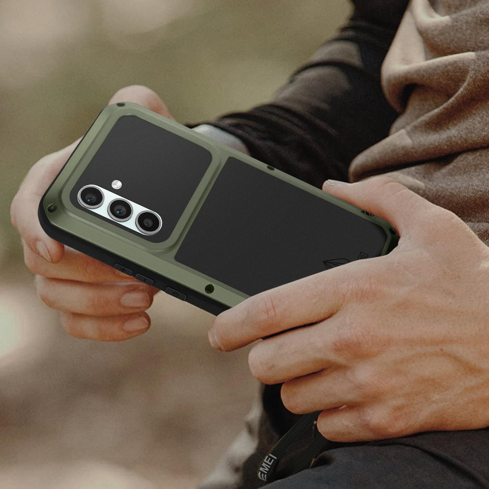 Mani di una persona che tengono un telefono con una custodia verde oliva. La fotocamera del telefono è visibile. Lo sfondo è sfocato.
