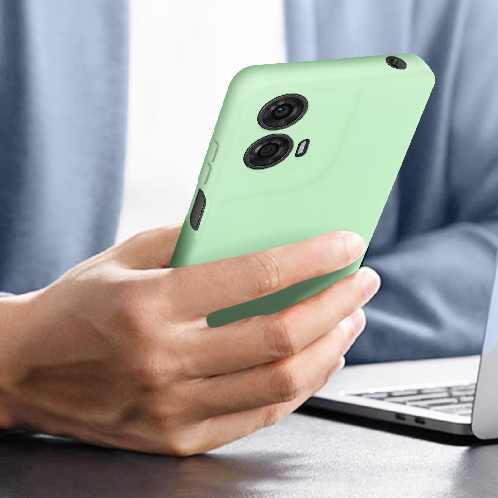 Eine Hand hält ein grünes Telefon. Das Telefon hat zwei Objektive. Im Hintergrund ein Laptop.