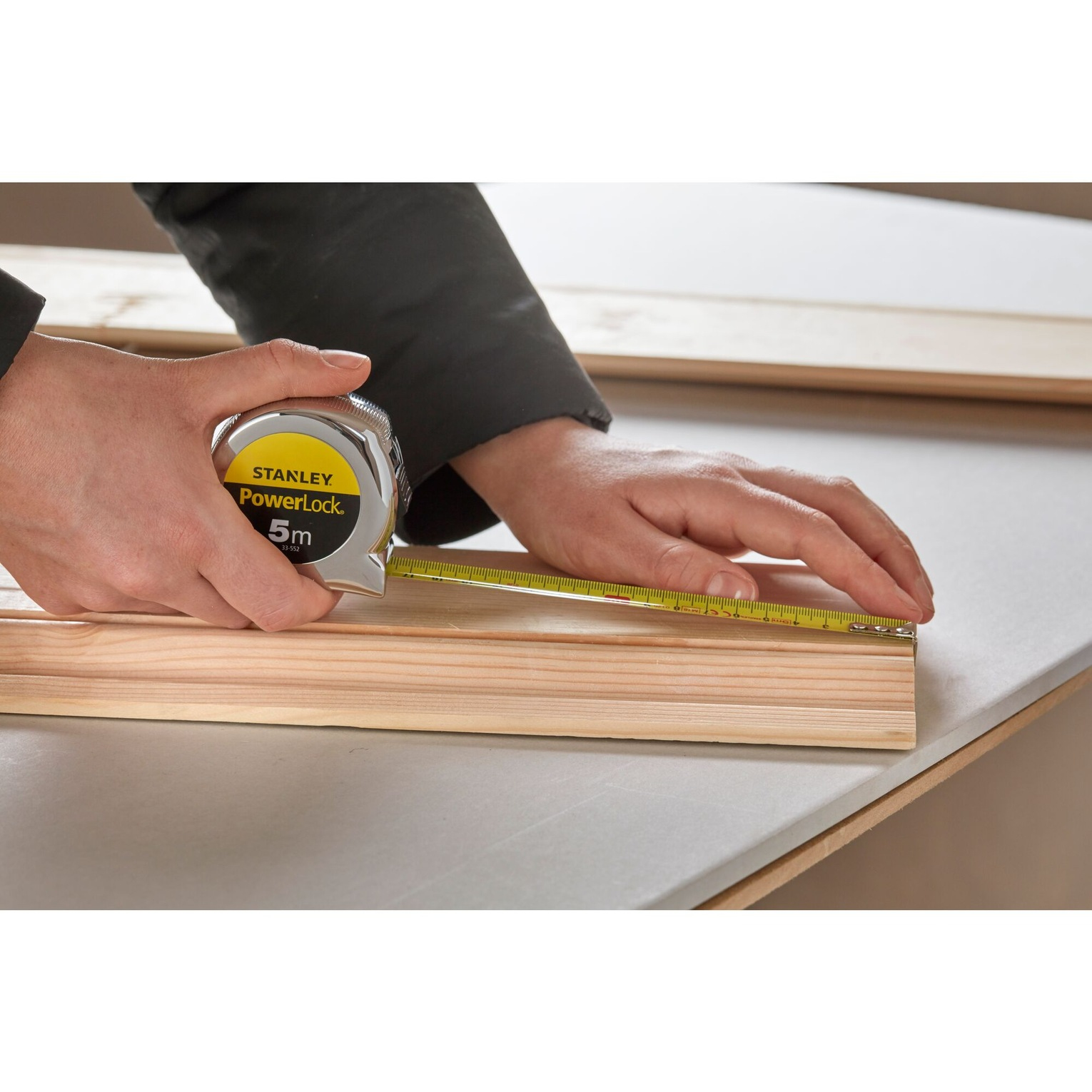 Osoba mierzy drewno za pomocą miarki. Drewno leży na szarej powierzchni.