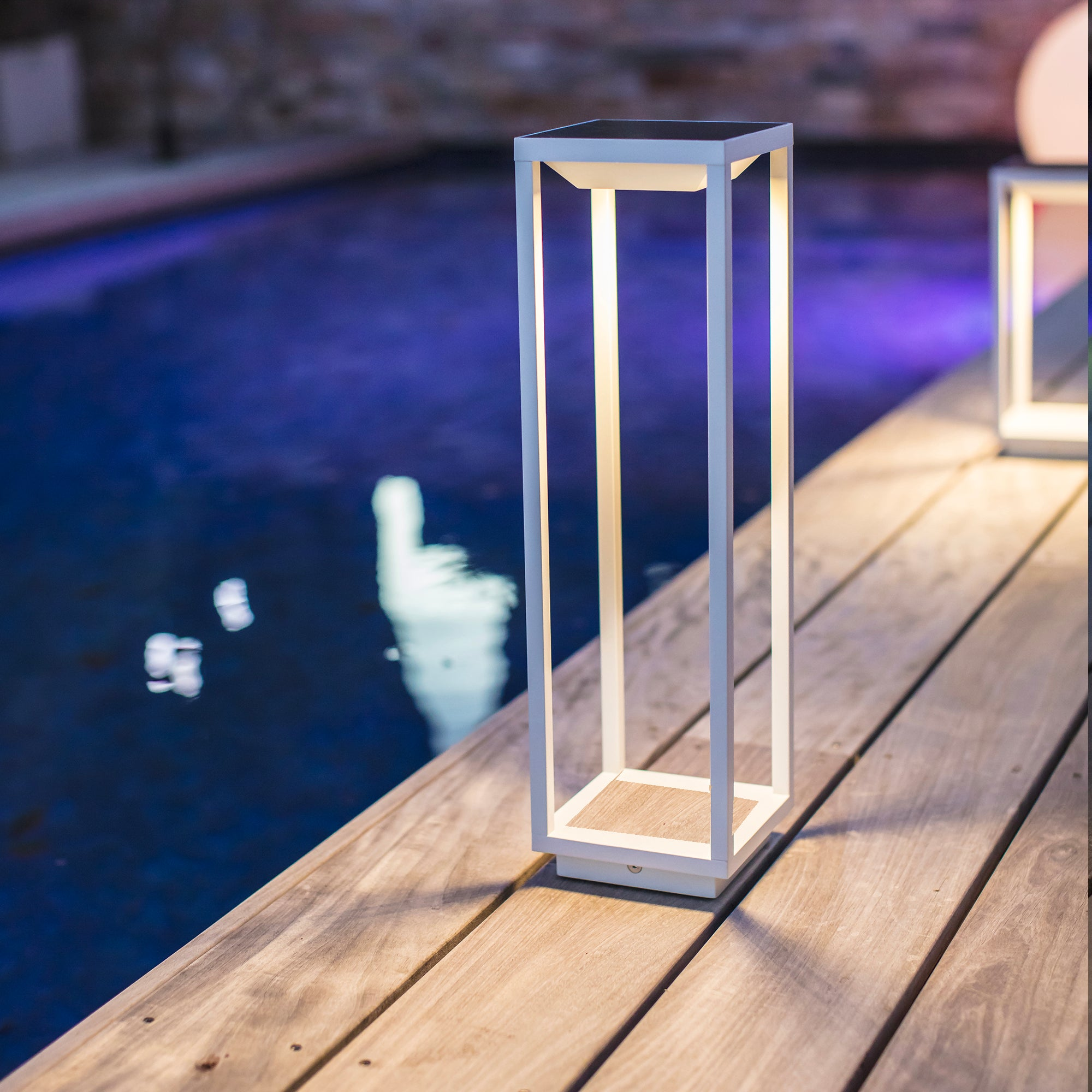 Eine weiße Außenleuchte auf einem Holzdeck neben einem Pool, mit blauem Wasser im Hintergrund.