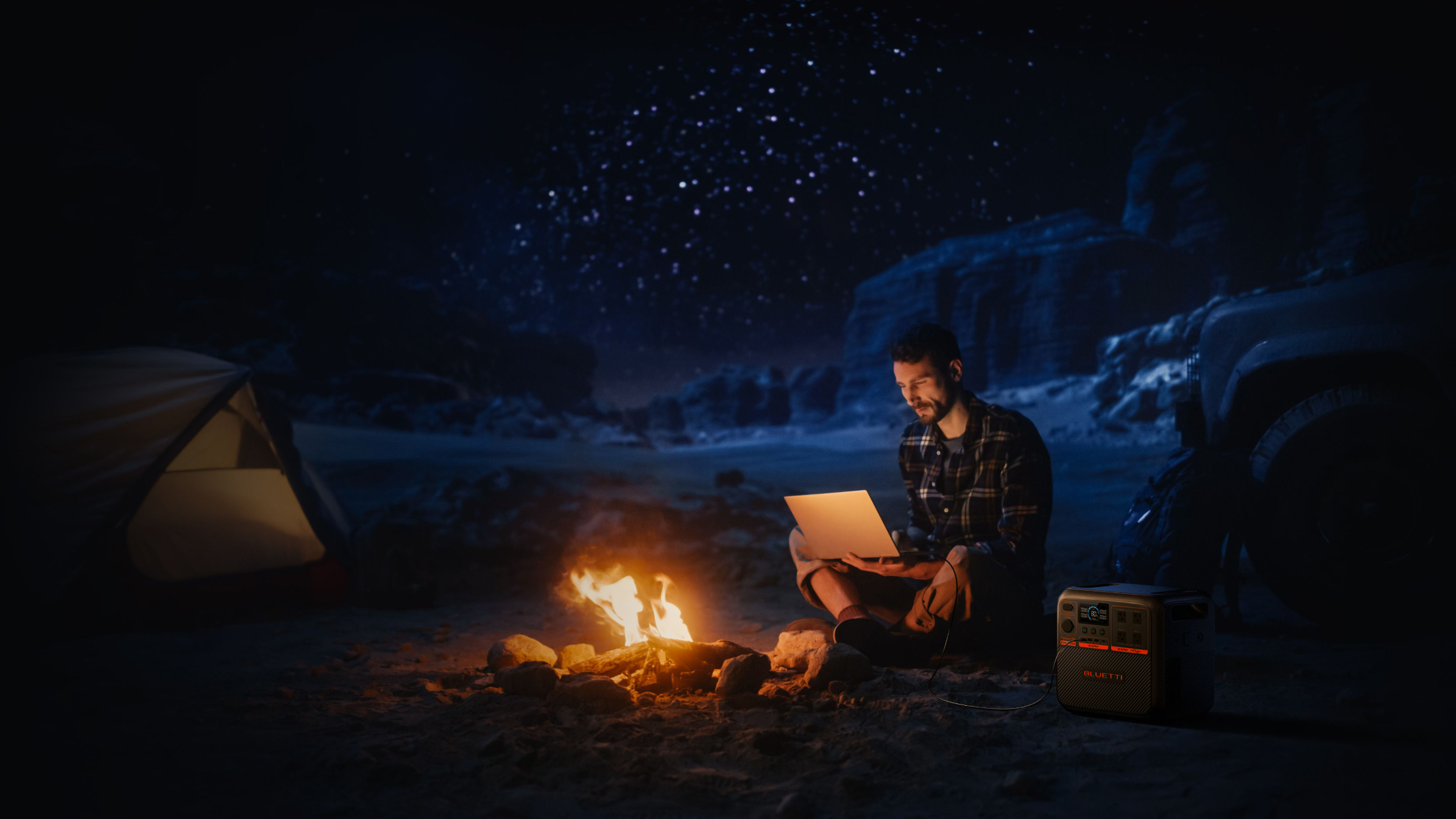 Mężczyzna używa laptopa przy ognisku w nocy. Namiot i samochód w tle.