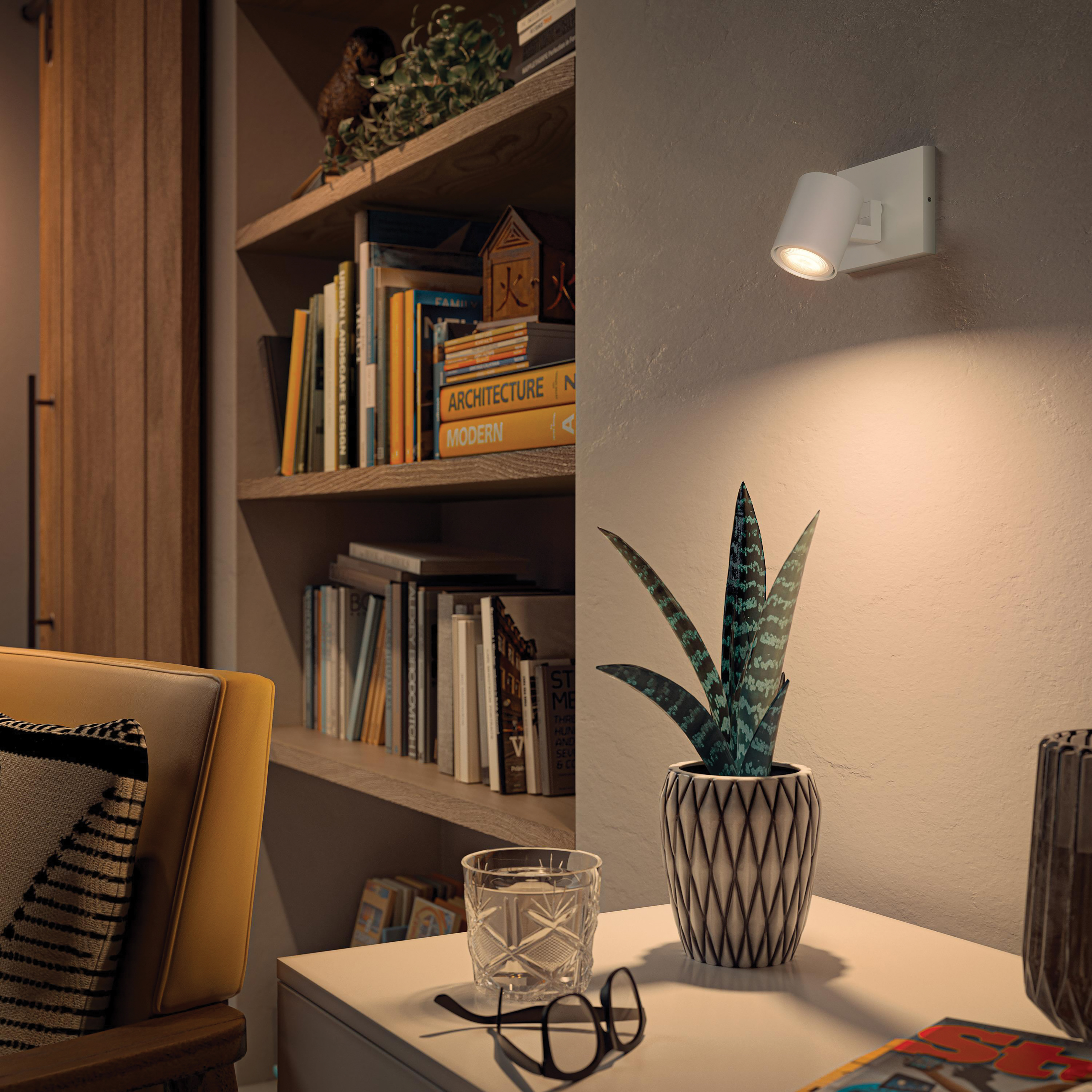 A well-lit bookshelf with books and decorative items, a table with a glass and glasses, and a wall lamp.