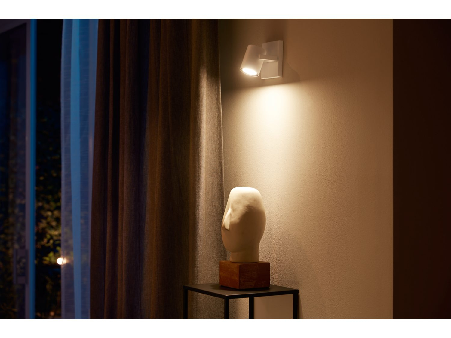 Białe światła na ścianie oświetlają rzeźbę. Zasłony i ciemne otoczenie w tle.