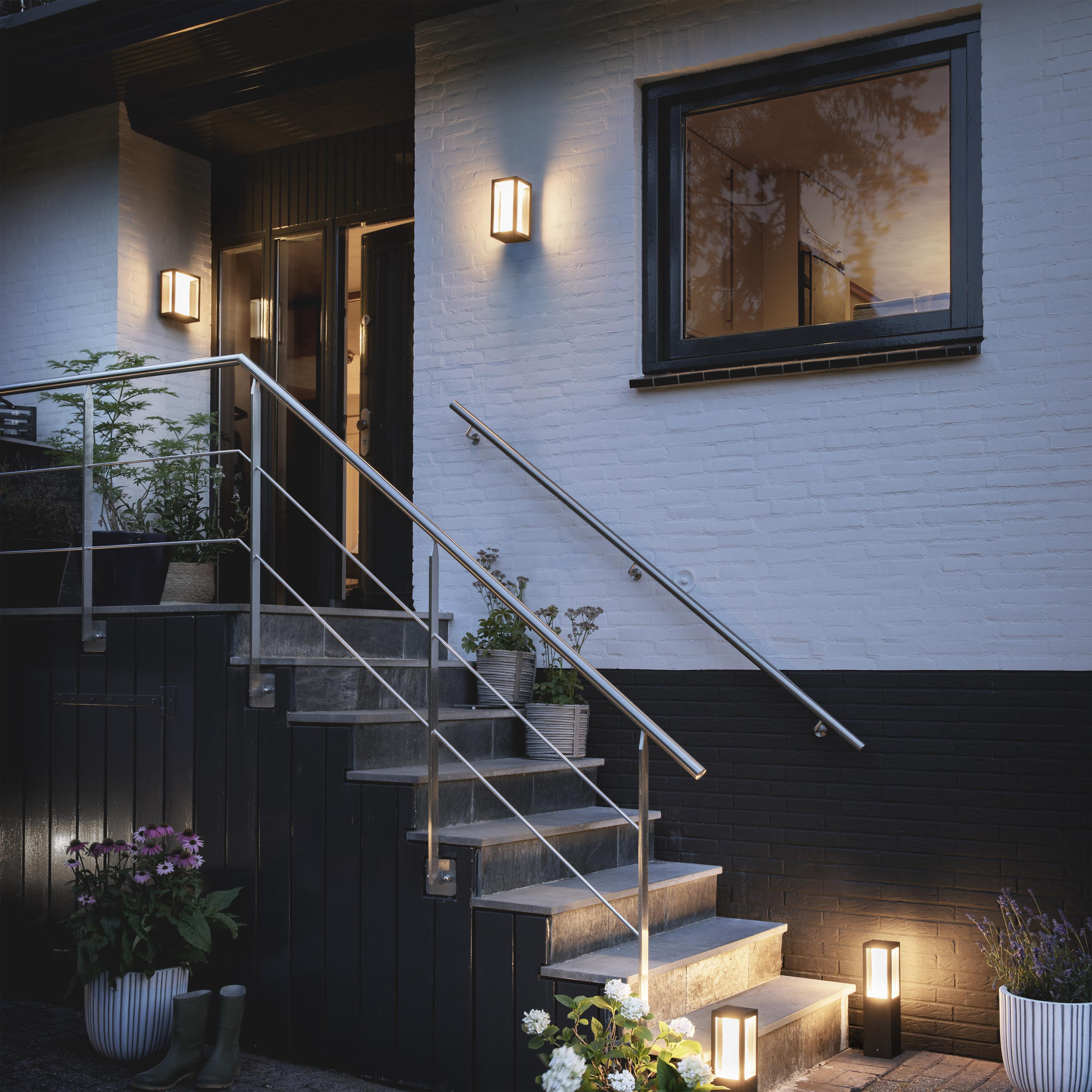 Extérieur d'une maison avec escalier, plantes et lumières. Le mur de briques contraste avec porte et fenêtre noires.
