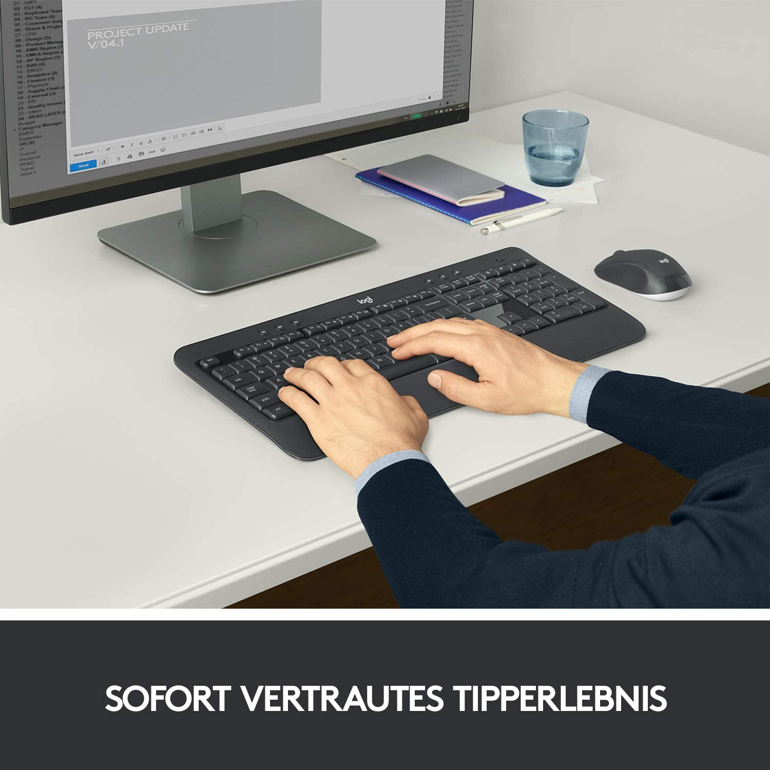 Osoba pisząca na czarnej klawiaturze. Monitor komputera i szklanka wody na biurku.