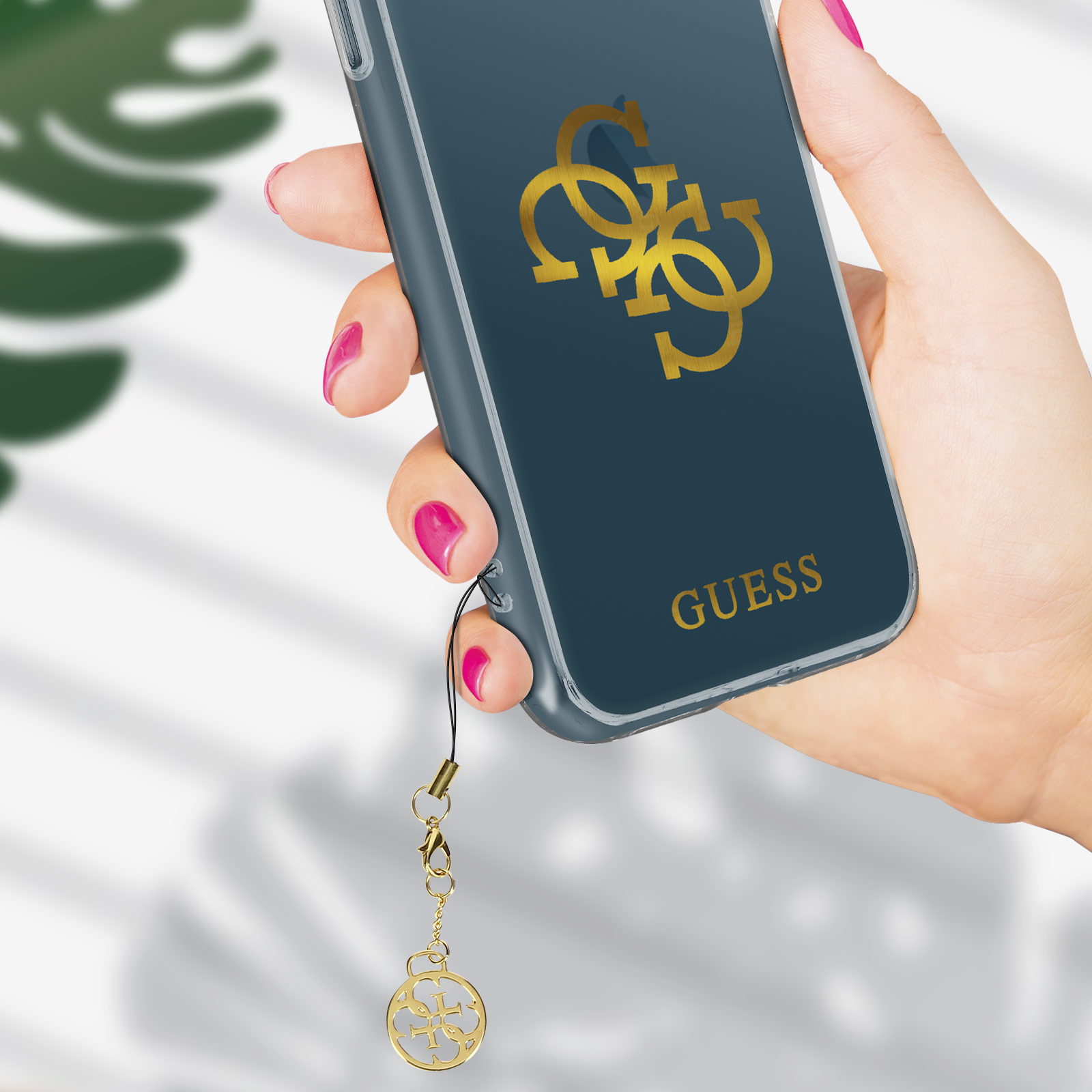 Mano sosteniendo teléfono con funda Guess. Logo dorado, fondo azul oscuro.