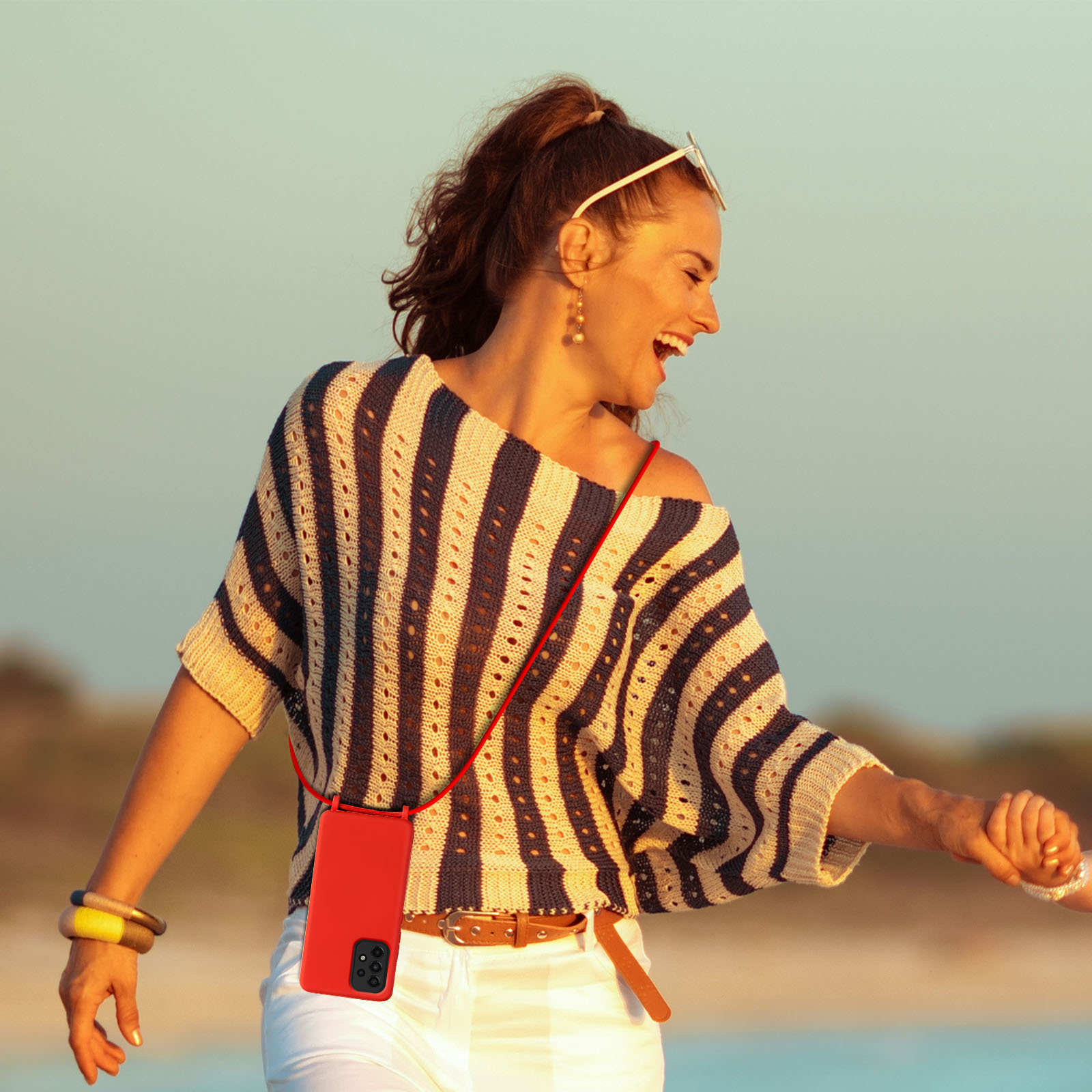 Une femme en haut rayé se tient la main, avec un étui rouge avec une lanière. Le fond est une plage.