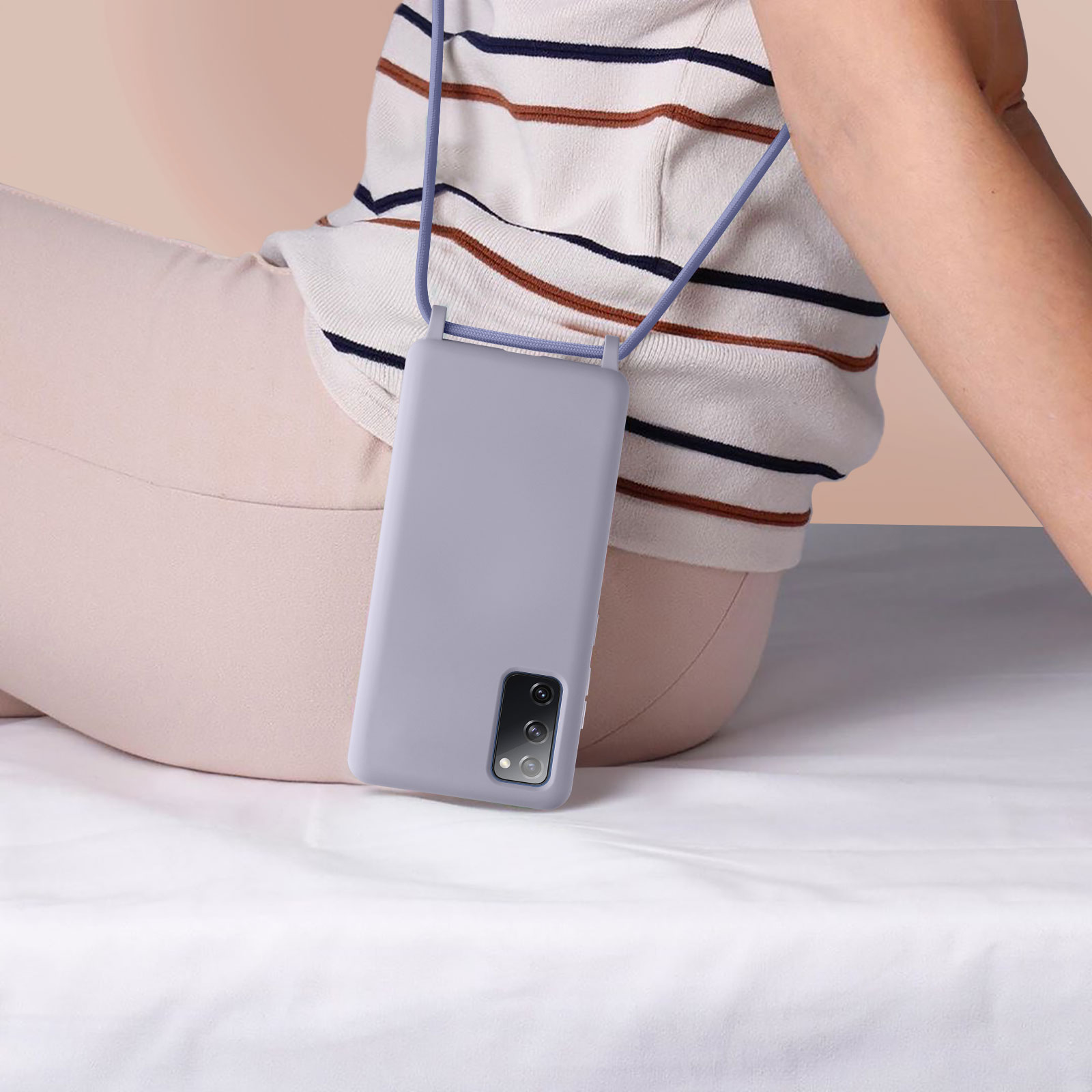 Personne tenant un téléphone lavande avec une lanière. Pantalon beige, chemise rayée, fond blanc.
