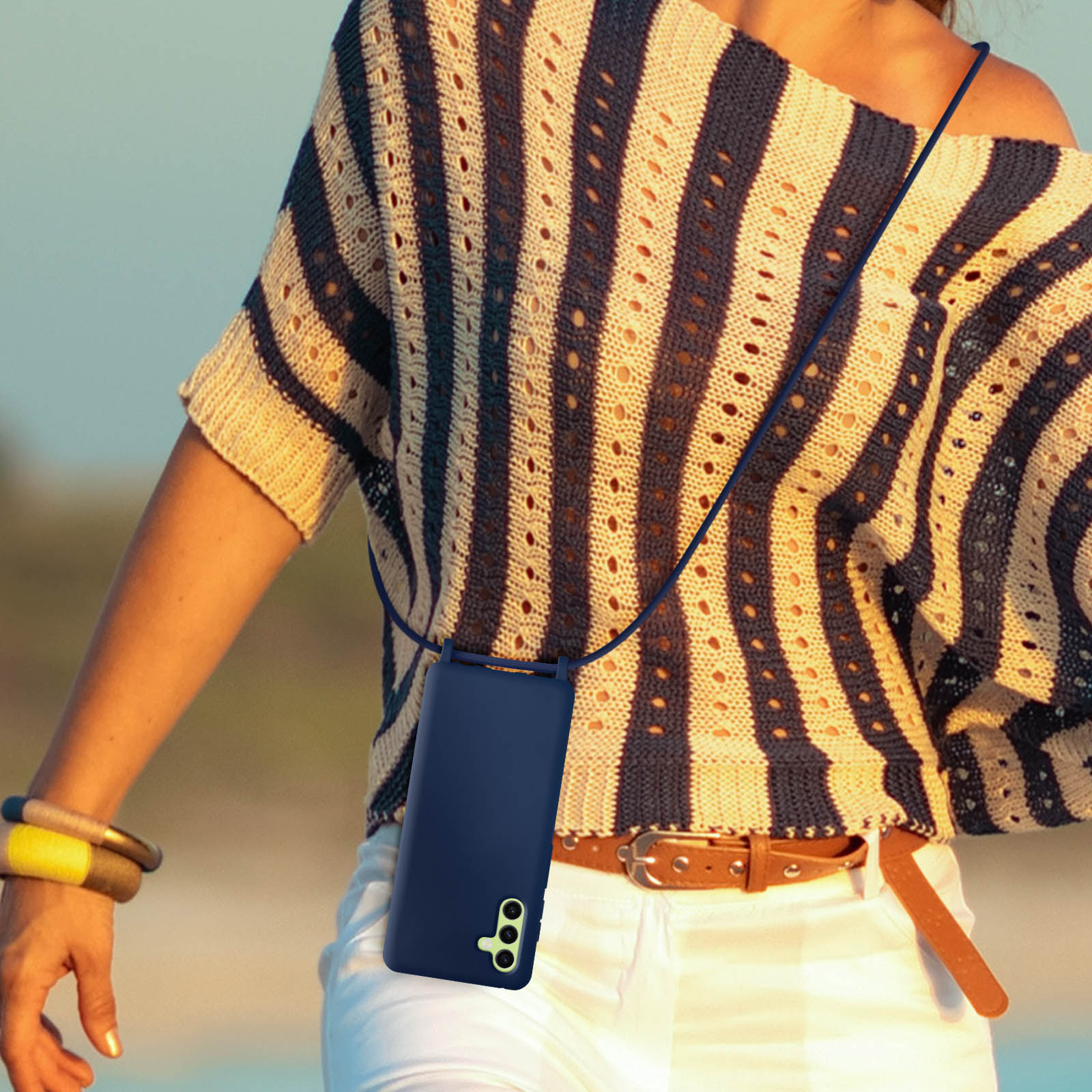 Une femme portant un haut rayé, un pantalon blanc, tenant un téléphone avec un étui bleu marine et une sangle.