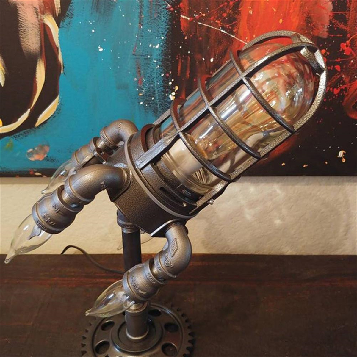 Tischlampe mit Metallkäfig um eine Glühbirne. Die Lampe hat rohrartige Arme. Der Hintergrund hat ein Gemälde mit Blau- und Rottönen.