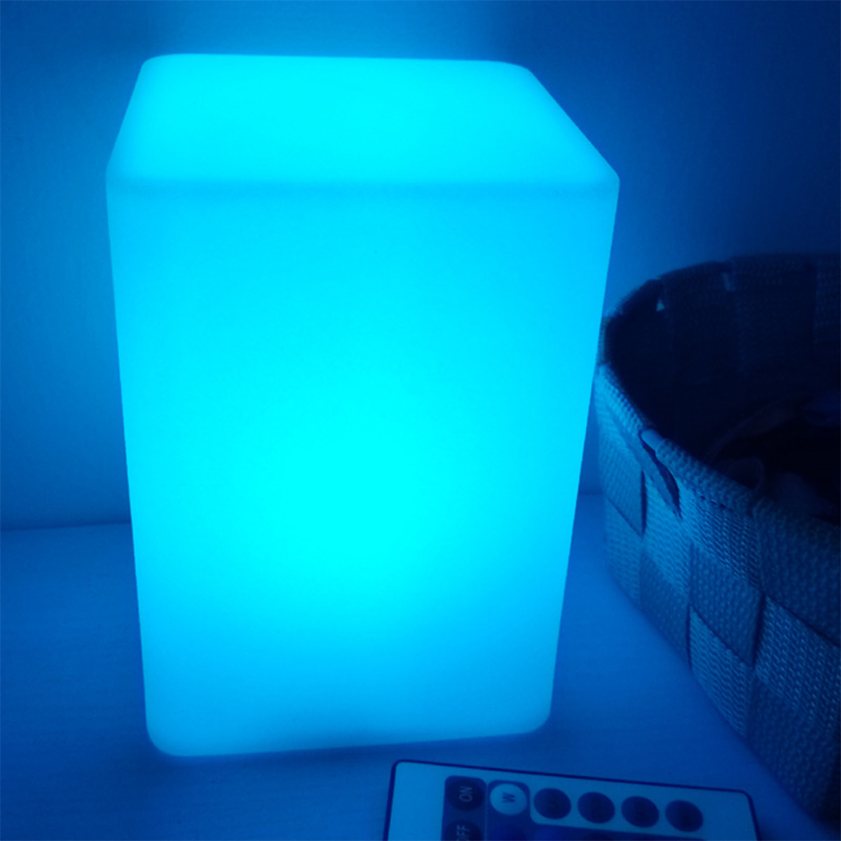 Eine blau leuchtende quadratische Lampe steht auf einem Tisch mit einem Korb und einer Fernbedienung.