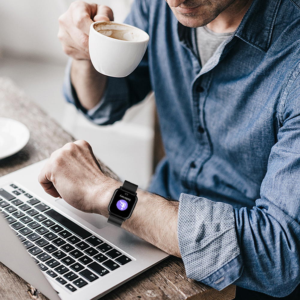 Ein Mann betrachtet eine Smartwatch an seinem Handgelenk, während er eine Kaffeetasse hält und einen Laptop benutzt.