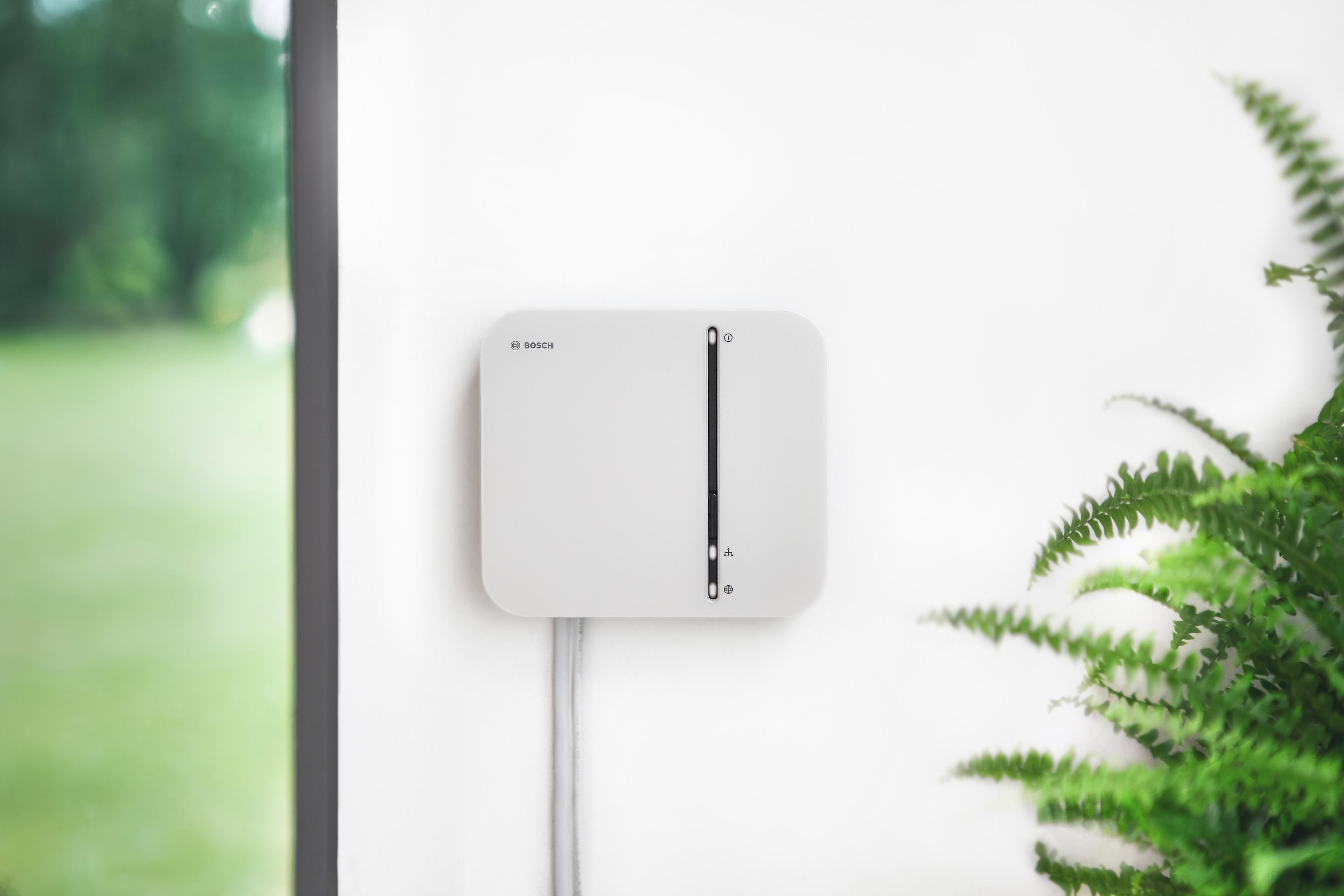 Ein weißes Bosch Smart Home Gerät an einer weißen Wand, mit einer grünen Pflanze in der Ecke.