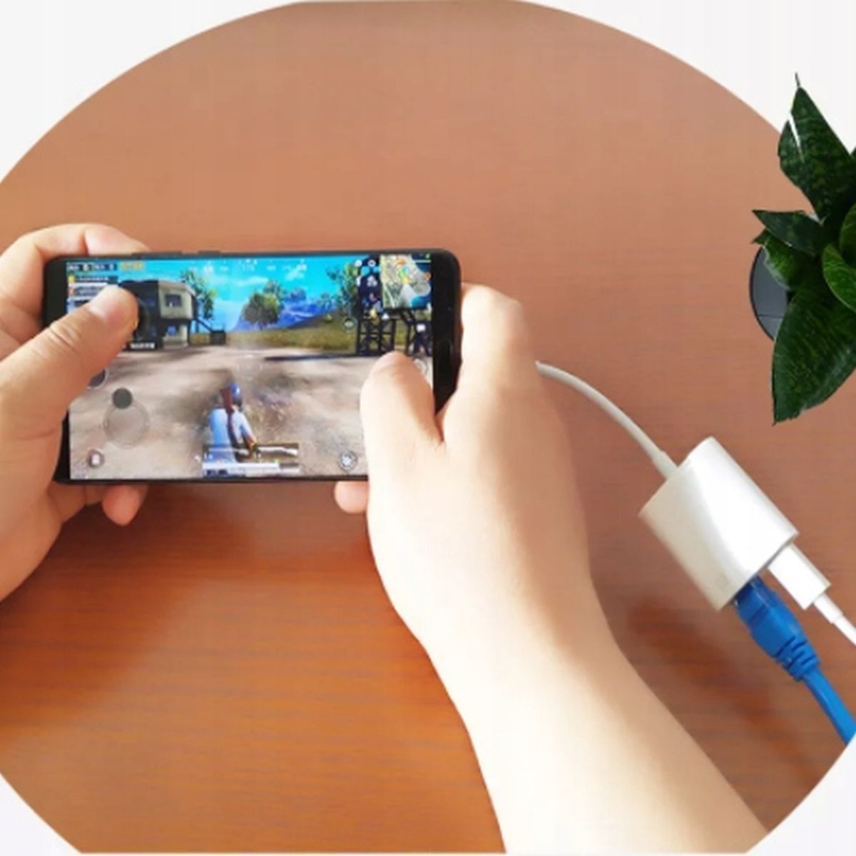 Ręce trzymające telefon z uruchomioną grą, podłączony do białej przejściówki z kablami.