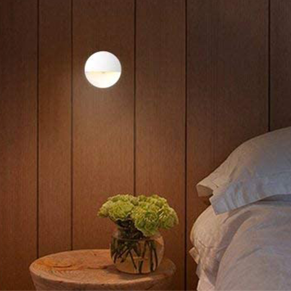 Runde weiße Wandleuchte beleuchtet einen Raum mit einer Vase grüner Blumen auf einem kleinen Holztisch.
