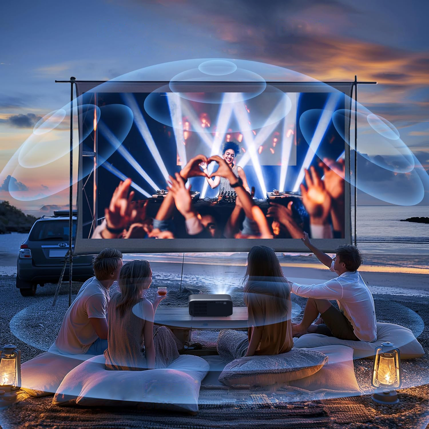 Ludzie oglądają film na dużym ekranie na zewnątrz, z samochodem i lampionami, o zmierzchu.