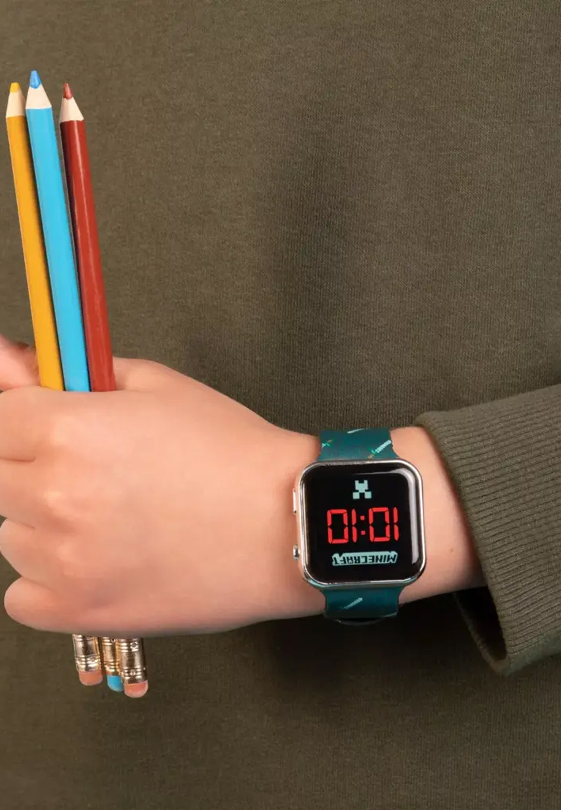 Personne portant une montre Minecraft et tenant des crayons de couleur. La montre affiche l'heure 10:01.