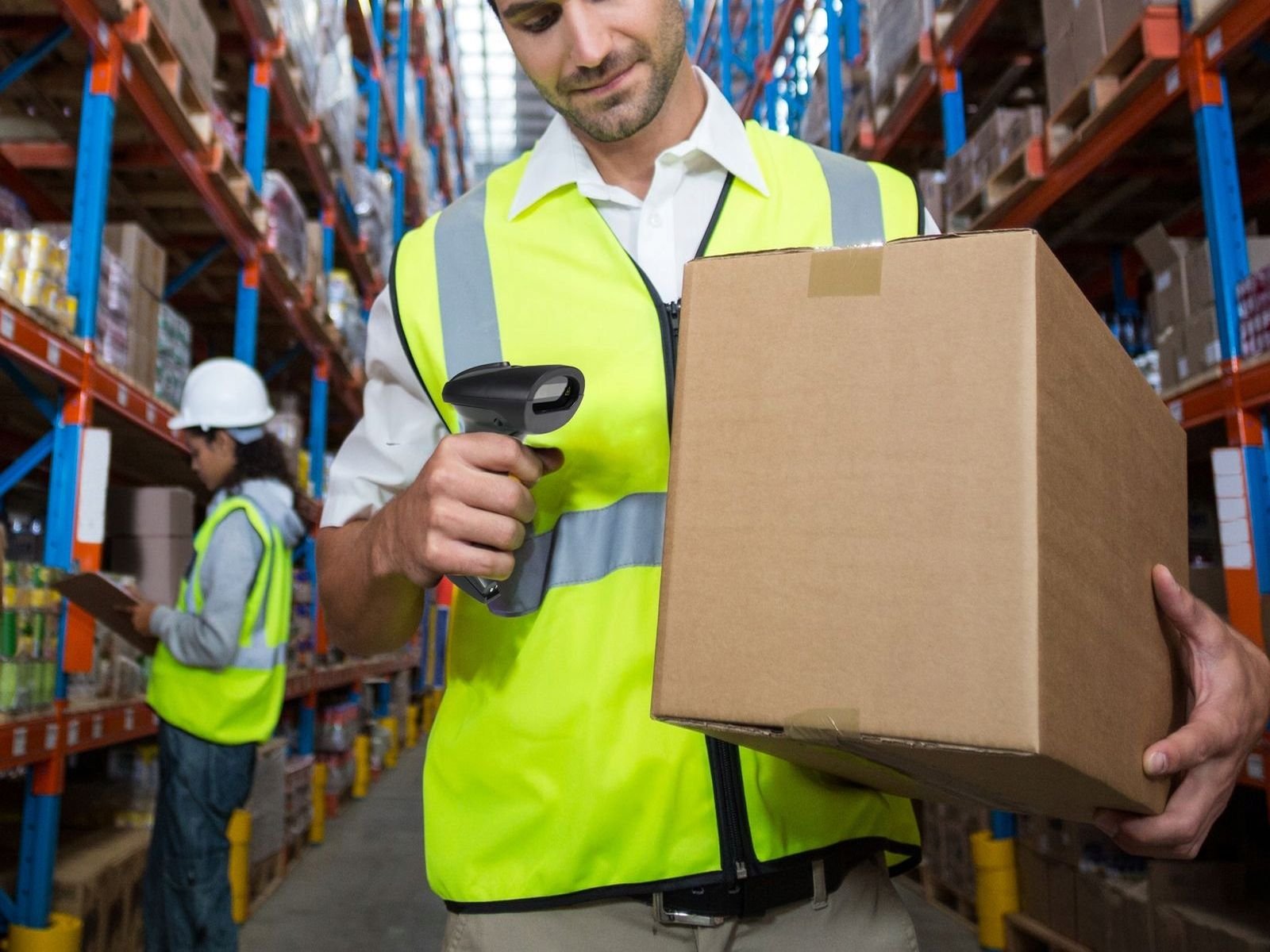 Mężczyzna w żółtej kamizelce skanuje pudełko w magazynie. Drugi pracownik jest w tle.