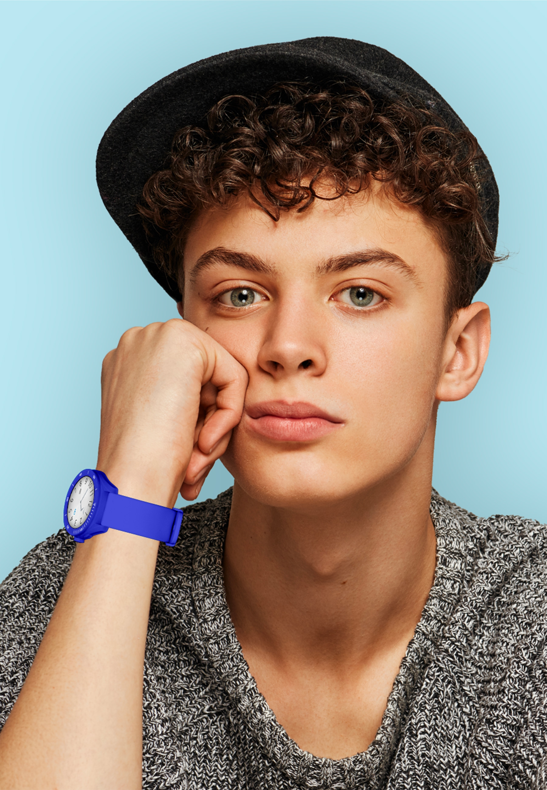 Jeune homme avec un chapeau, portant une montre à bracelet bleu. Il s'appuie sur sa main, regardant la caméra.